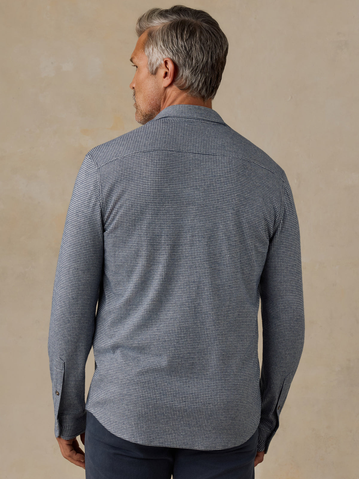 A man viewed from the back wearing a long-sleeve button-down shirt with a fine micro-check pattern in gray and white, paired with dark trousers, set against a textured beige background., image 3 of 7