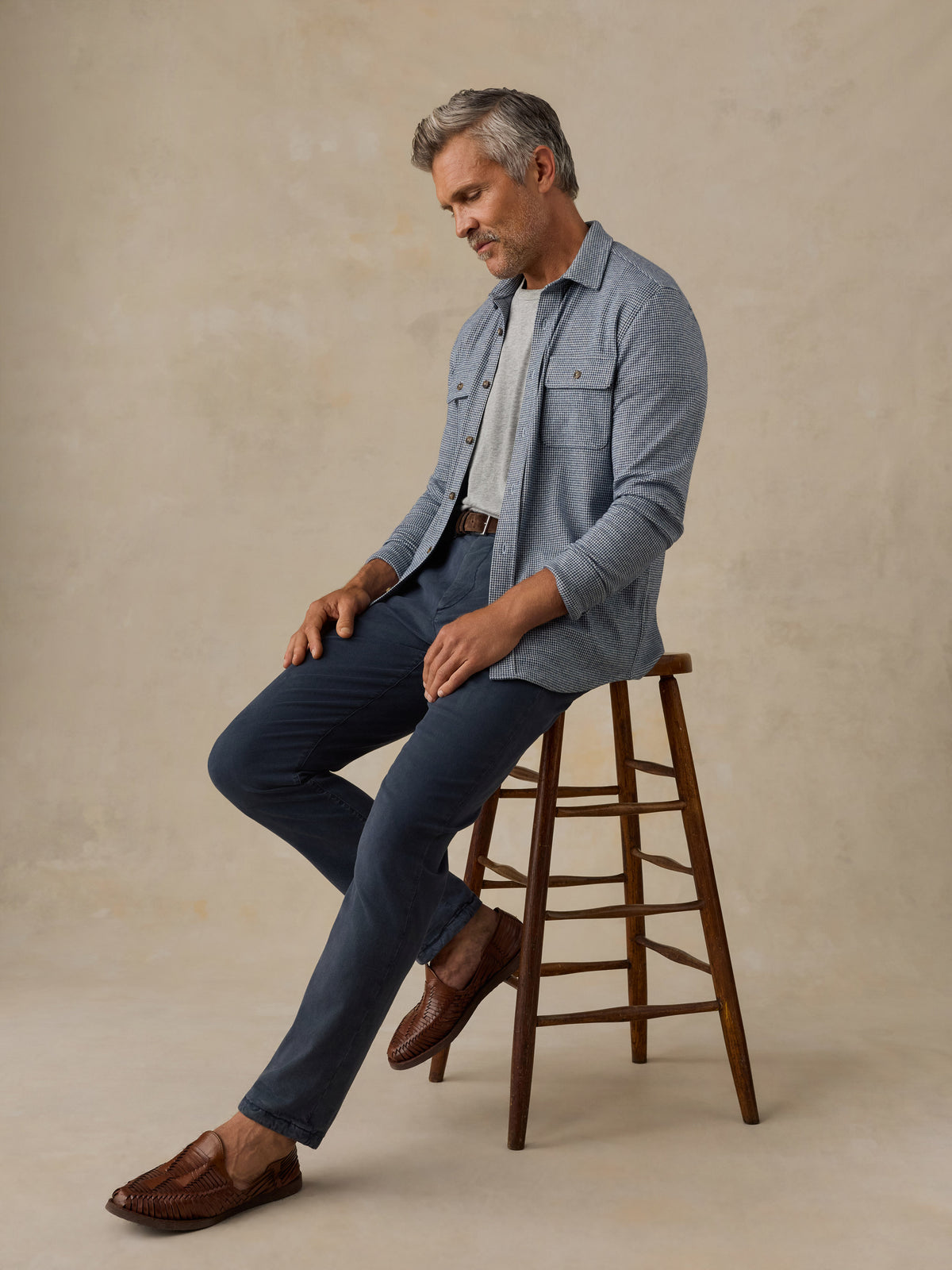 A man with gray hair sits on a wooden stool wearing a light blue denim shirt over a white t-shirt, navy blue pants, and brown leather loafers, against a textured beige studio backdrop., image 4 of 7
