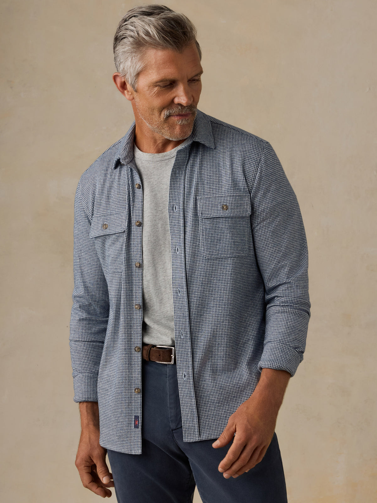 A man wearing a light blue and white micro-check patterned button-down shirt over a light gray t-shirt, paired with navy pants and a brown belt, standing against a neutral beige background., image 2 of 7