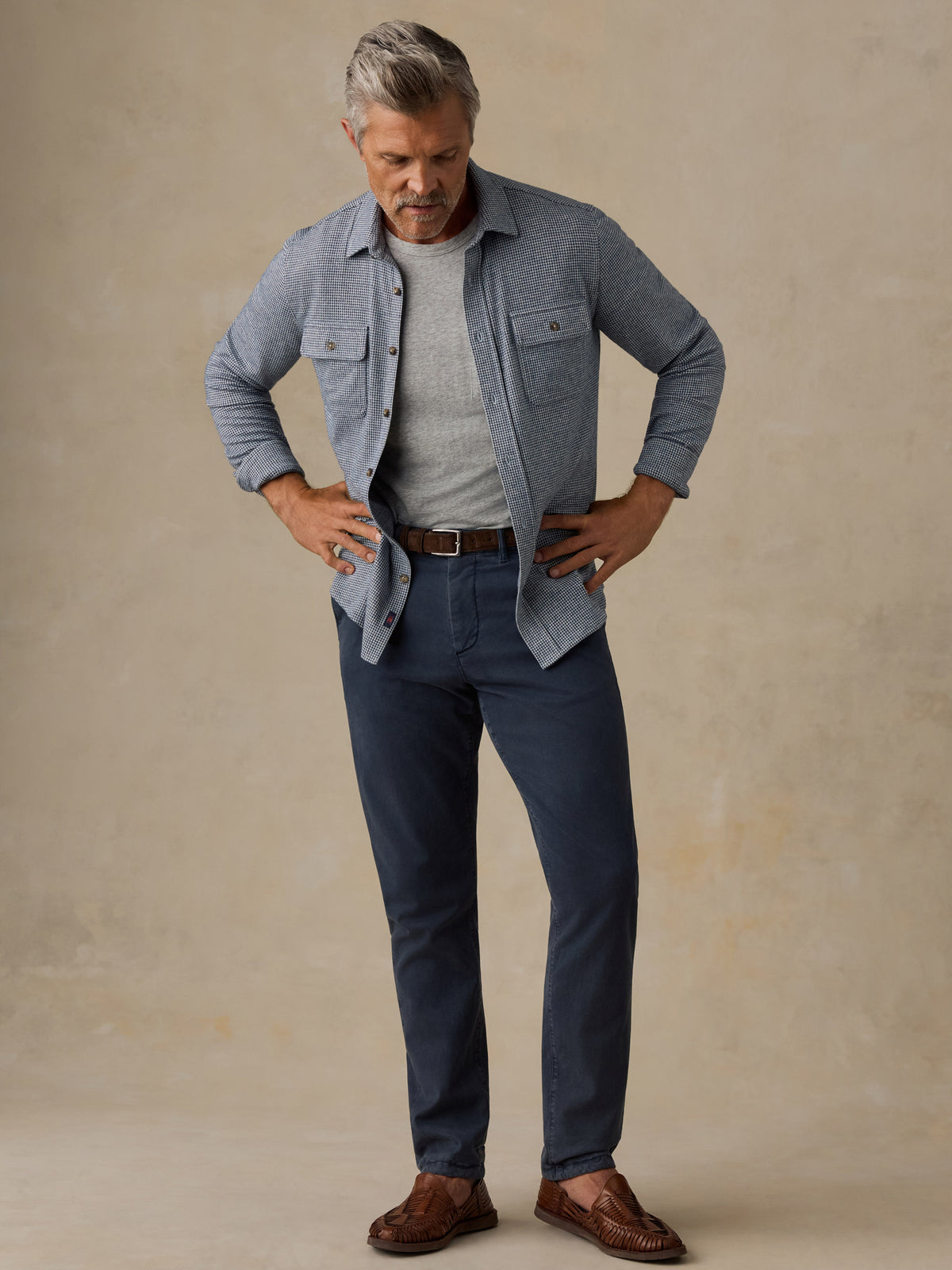 A man modeling a light blue button-up shirt over a gray t-shirt, paired with navy blue jeans, a brown belt, and brown leather loafers, standing against a textured beige studio backdrop., image 7 of 7
