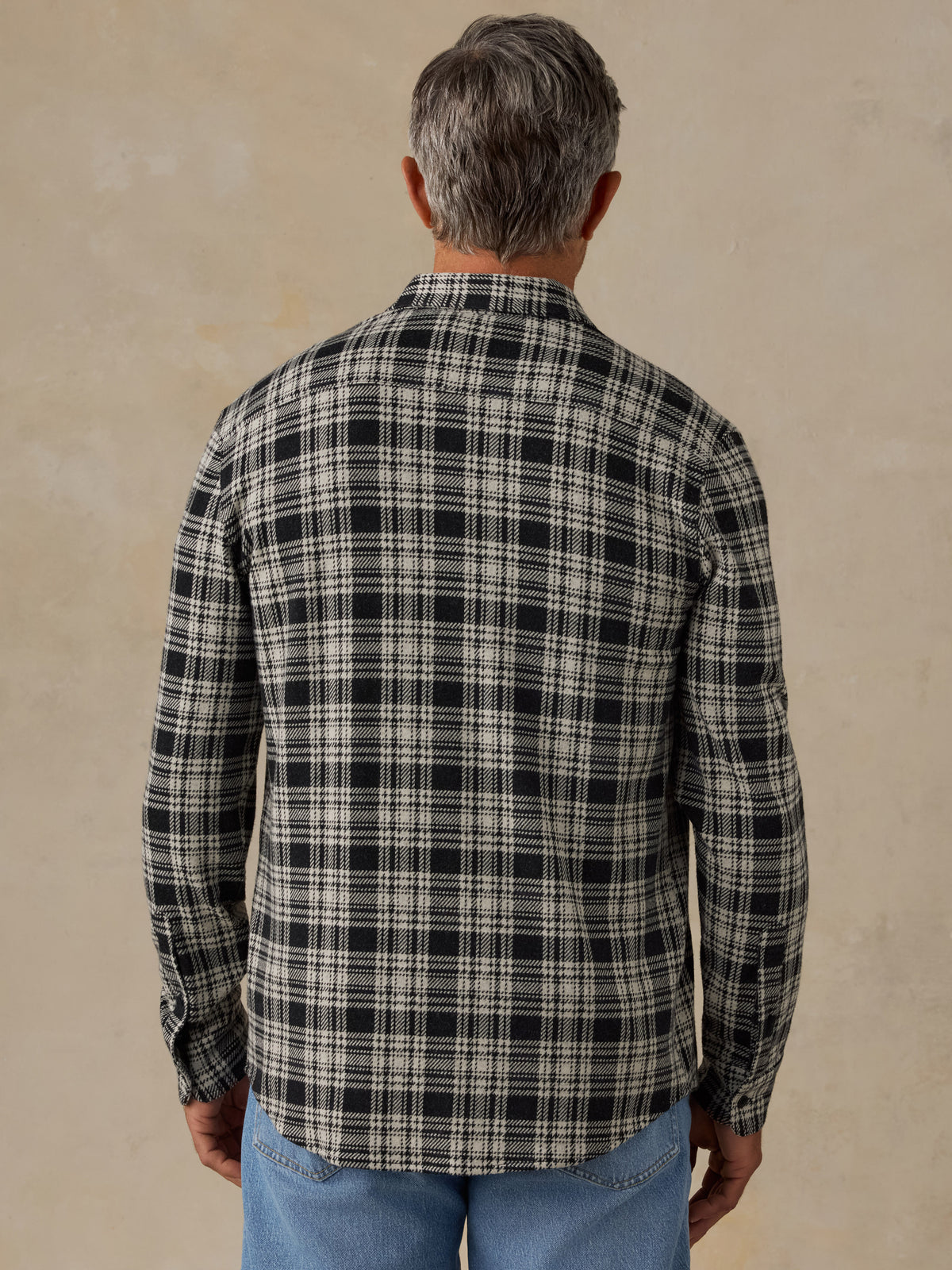 A man viewed from behind wearing a long-sleeve plaid button-up shirt in black and off-white, paired with blue jeans, against a textured beige background., image 3 of 6