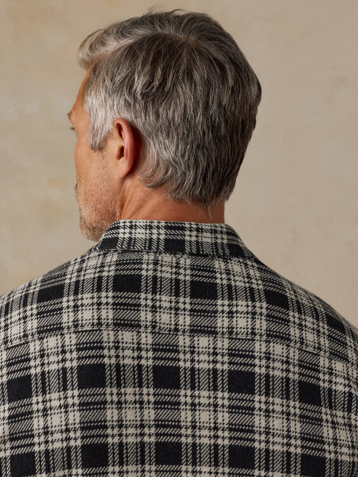 Back view of a man with gray hair wearing a black and off-white plaid flannel shirt against a neutral beige background., image 6 of 6