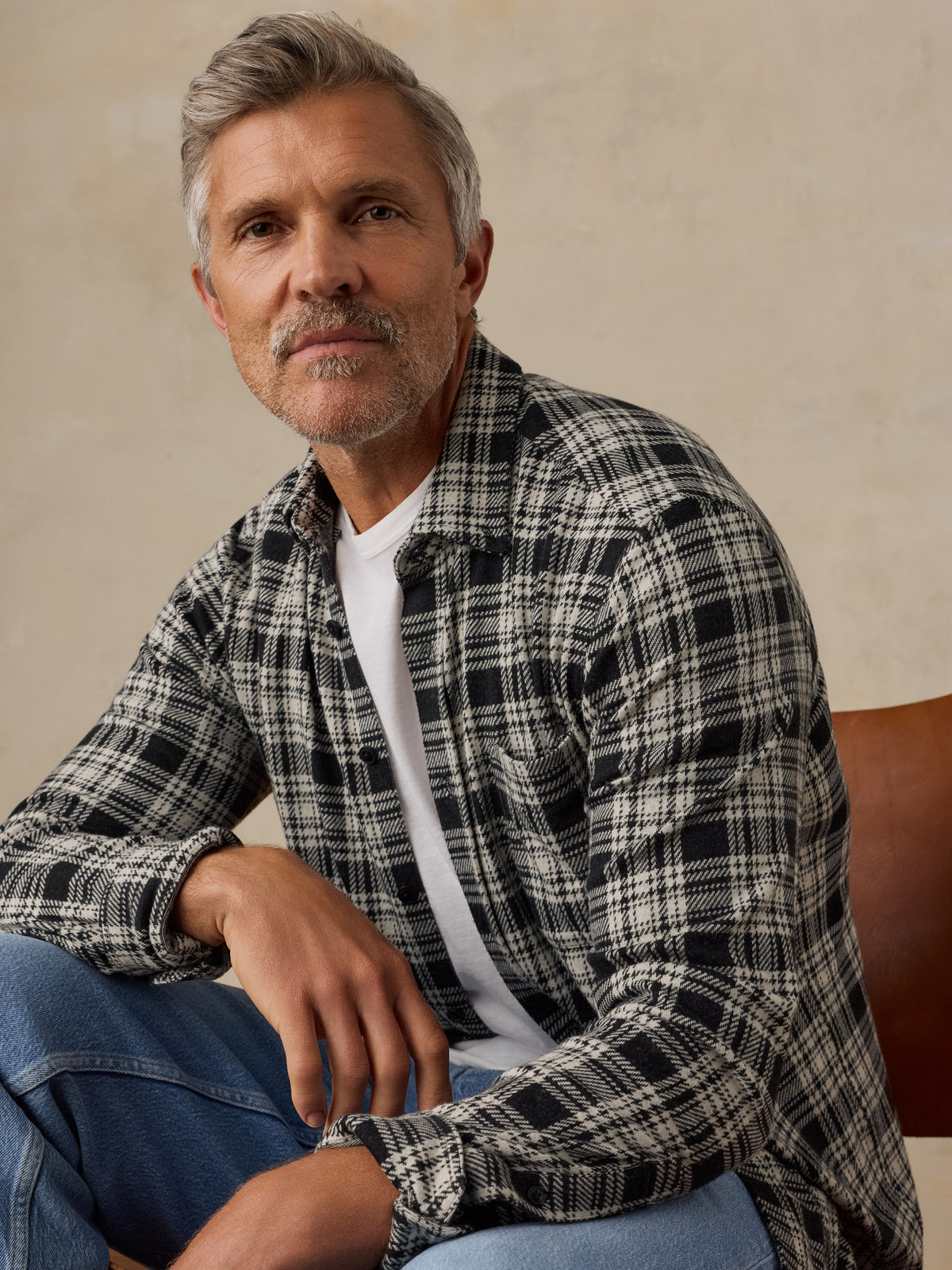 A man with gray hair and a beard wearing a black and white plaid button-up shirt over a white t-shirt and blue jeans, sitting in a leather chair against a textured beige wall., image 5 of 6