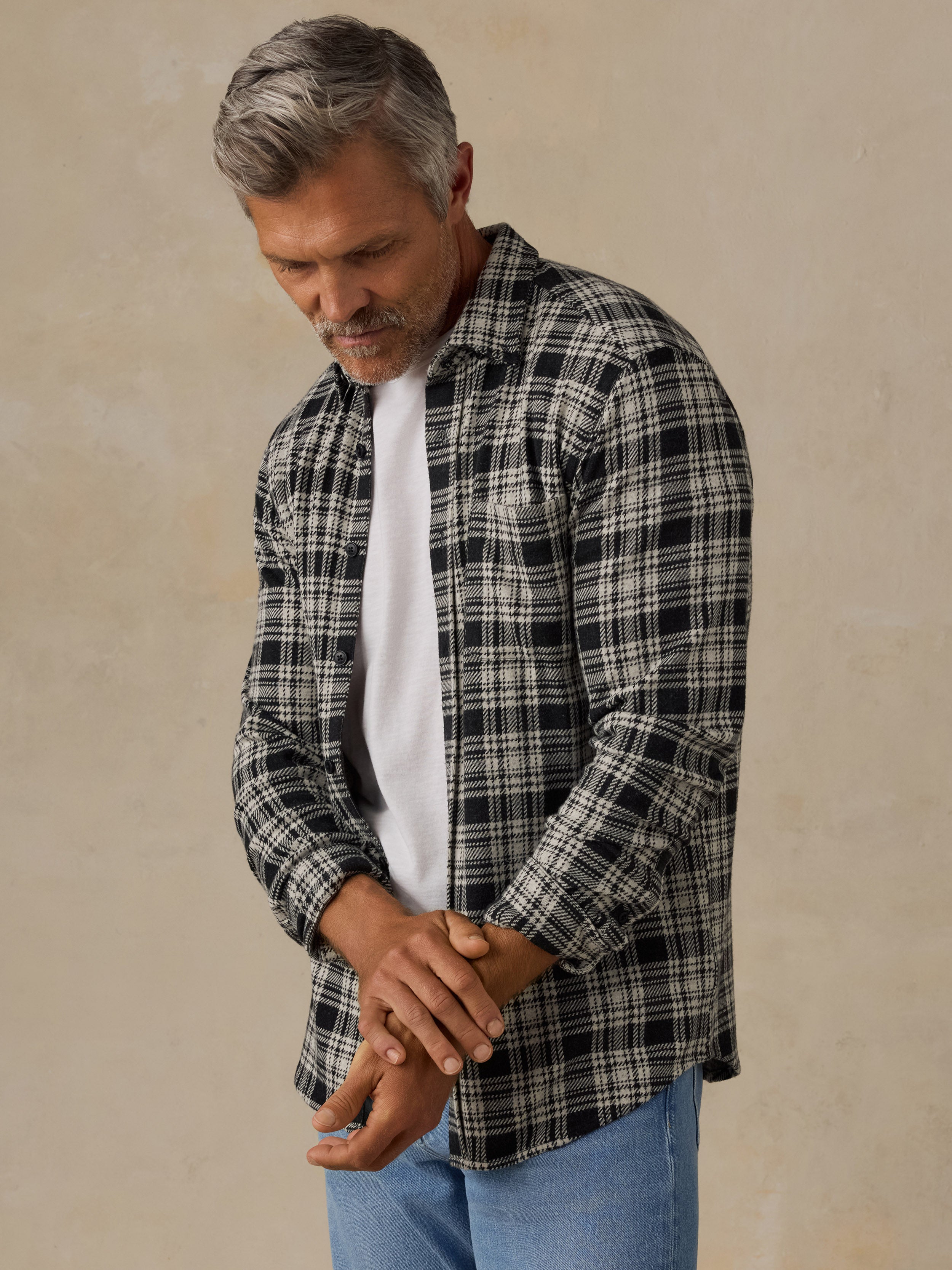 A man wearing a black and white plaid button-down shirt over a white t-shirt and blue jeans, standing against a textured beige wall.
