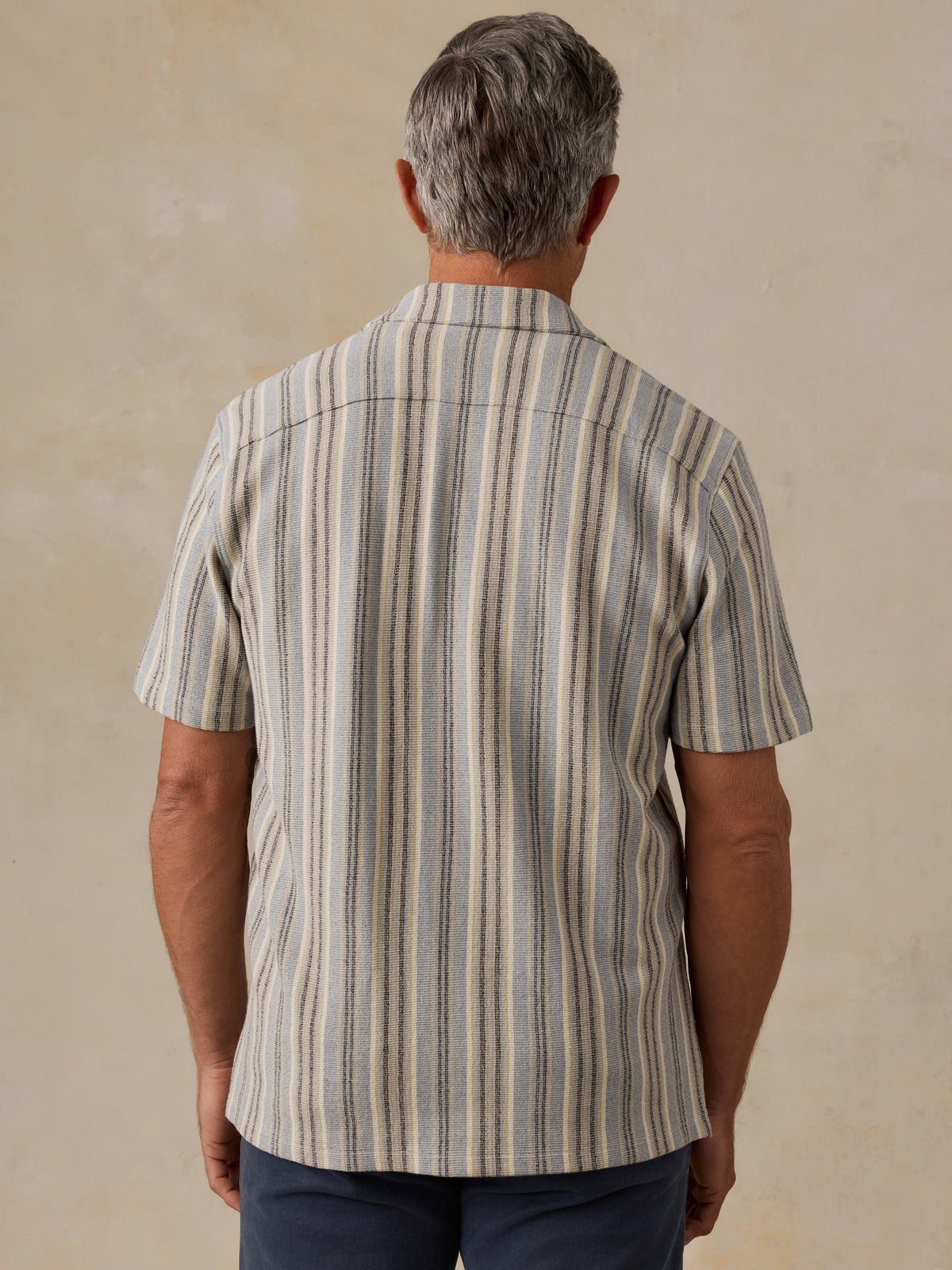 A man viewed from behind wearing a short-sleeve button-up shirt with vertical stripes in beige, gray, and brown tones, paired with dark blue pants, set against a textured beige background., image 3 of 6