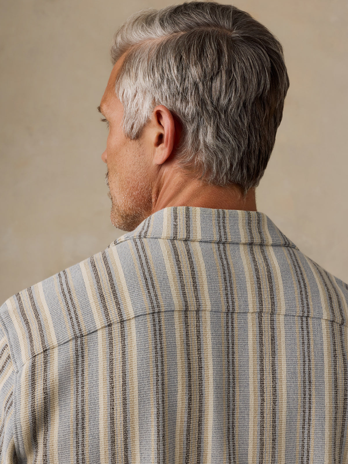 A close-up back view of a man with gray hair wearing a textured striped button-down shirt in beige, gray, and navy blue., image 6 of 6