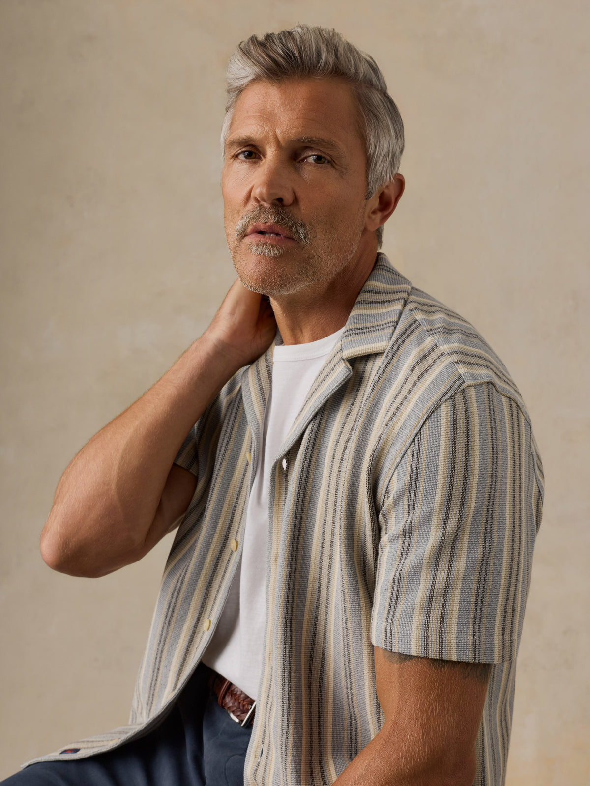 A man with gray hair and a mustache wearing a short-sleeve button-up shirt with vertical beige, gray, and white stripes over a white t-shirt, posed against a neutral textured beige background., image 5 of 6