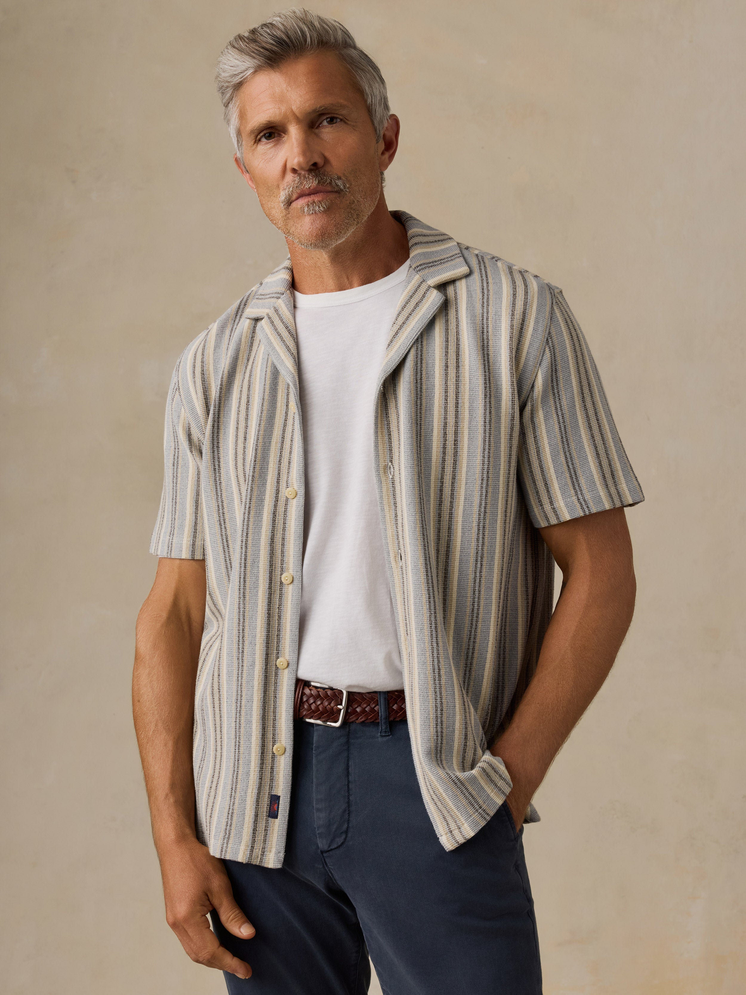 A man wearing a short-sleeve button-up shirt with vertical beige, gray, and blue stripes over a white t-shirt and navy pants, standing against a textured beige background.