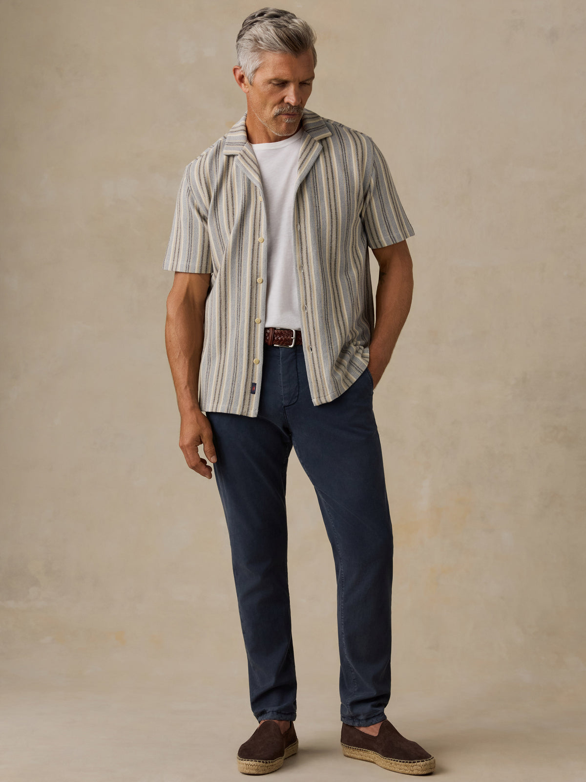 A man wearing a beige and gray vertically striped short-sleeve button-up shirt over a white t-shirt, paired with navy blue jeans and brown slip-on shoes, standing against a textured beige studio backdrop., image 4 of 6