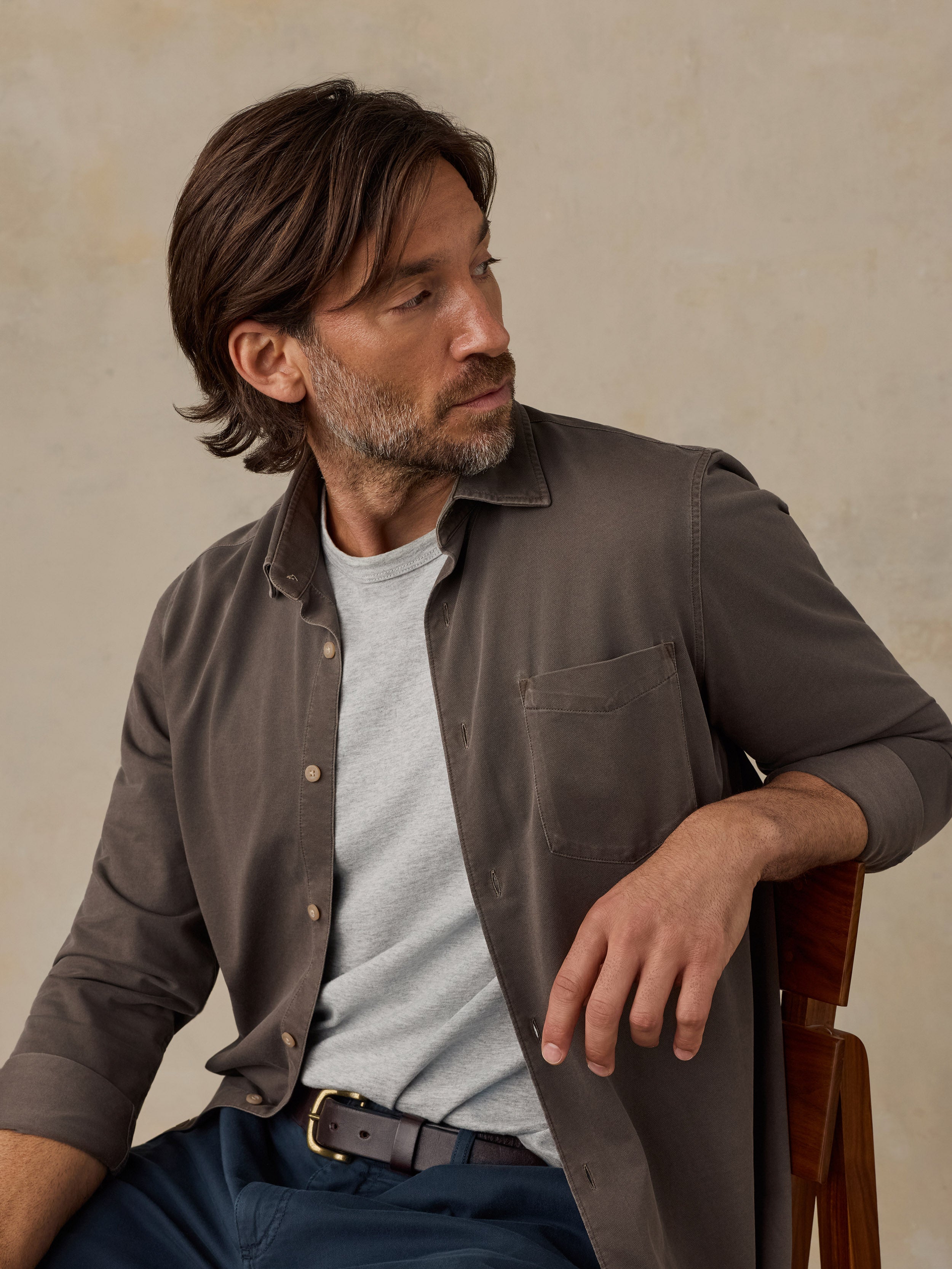 Man sitting on a wooden chair wearing a brown button down shirt and navy chino pants against a beige background