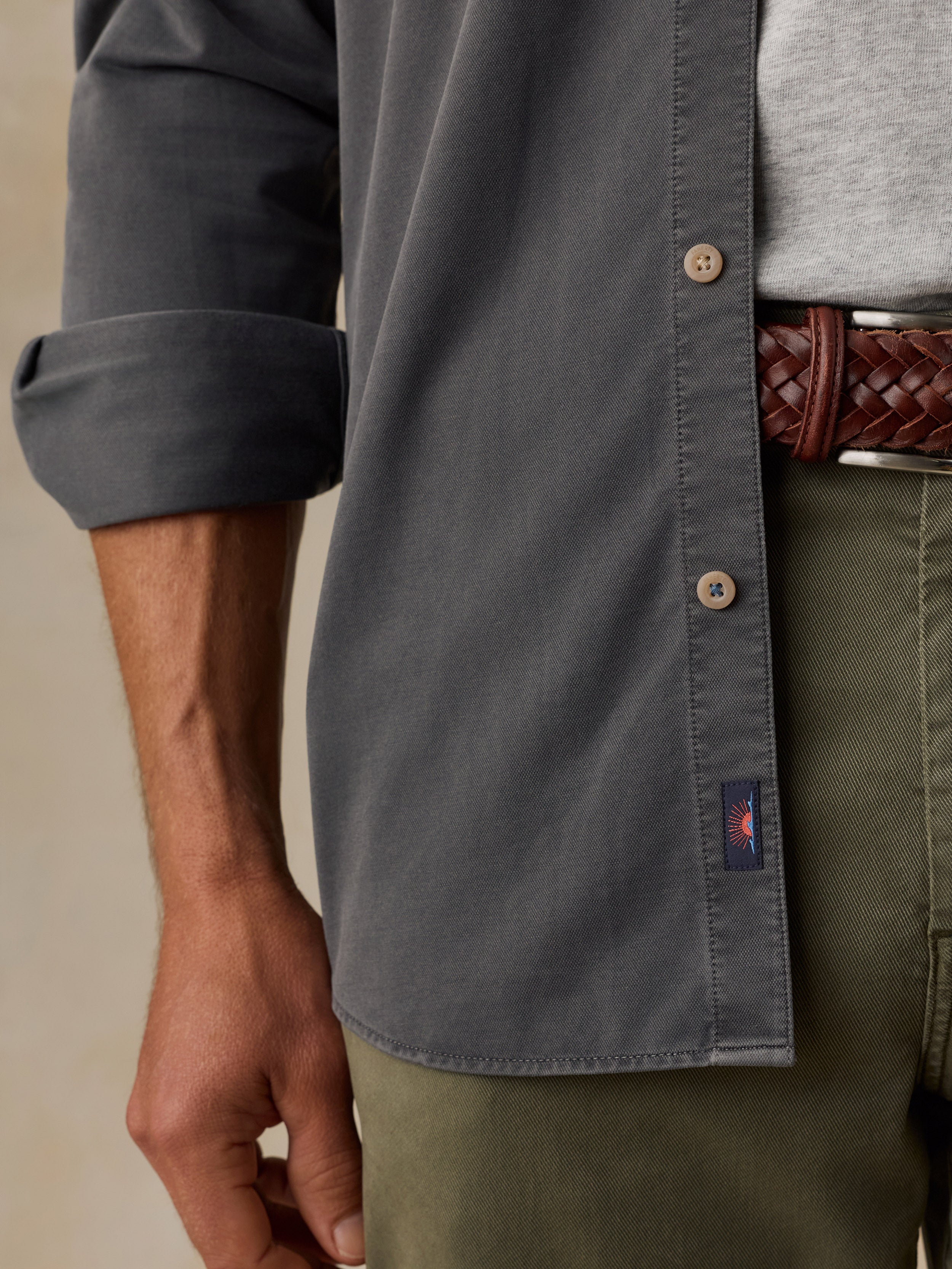 Close-up of a man wearing a dark gray long-sleeve button-down shirt with rolled sleeves, paired with olive green trousers and a brown braided belt.