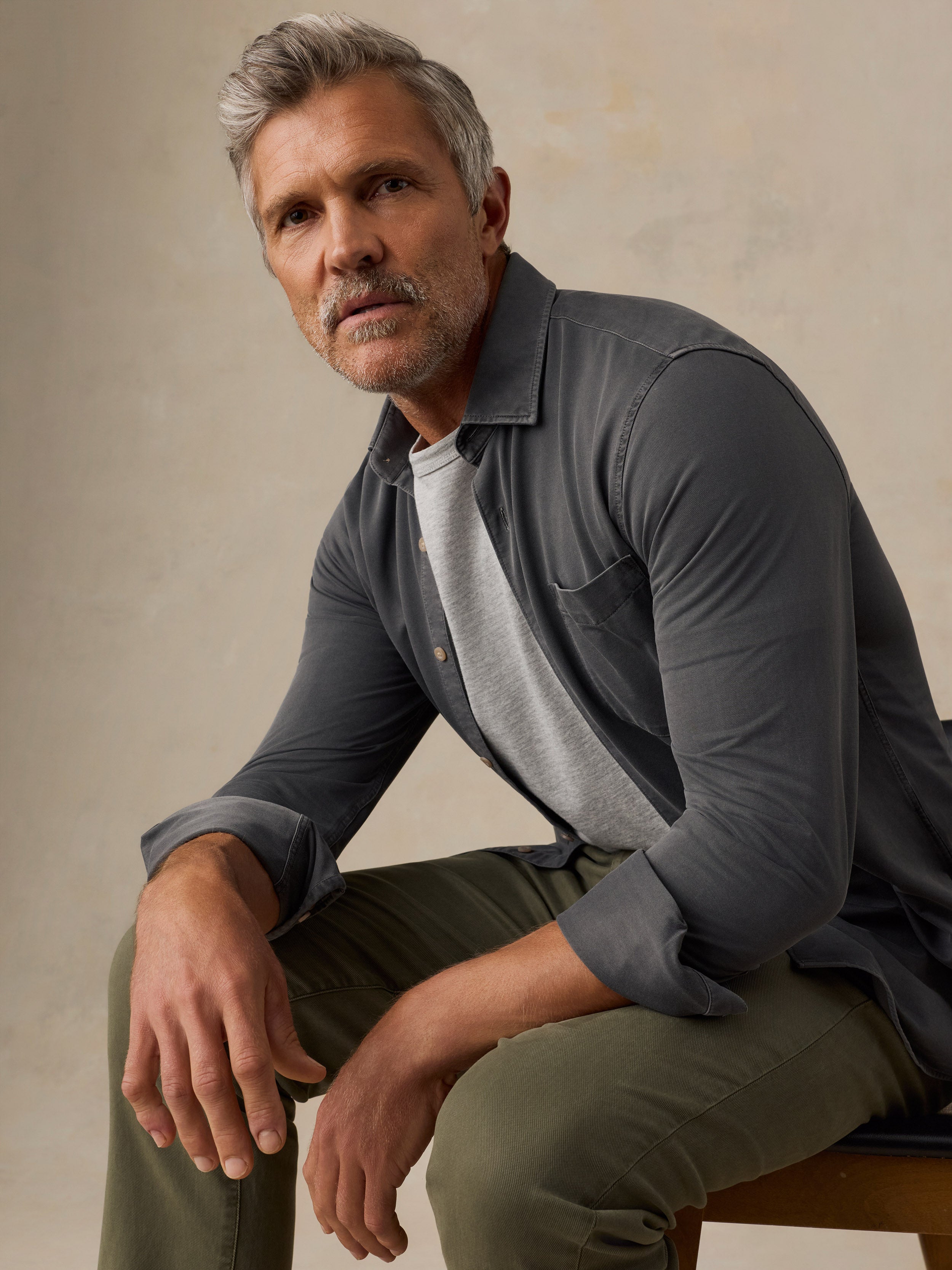 A man with gray hair and a mustache sits on a stool wearing a dark gray long-sleeve button-up shirt over a light gray t-shirt and olive green pants, against a textured beige studio background.