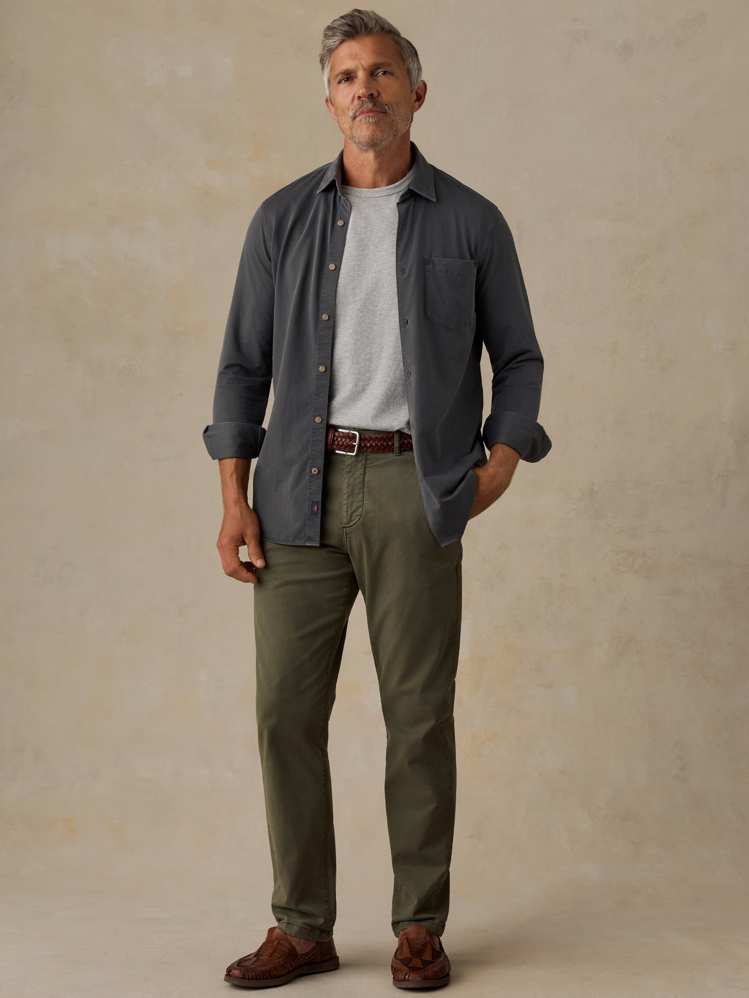 A man wearing a dark slate blue button-down shirt over a white t-shirt, olive green pants, and brown leather shoes, standing against a textured beige studio backdrop.