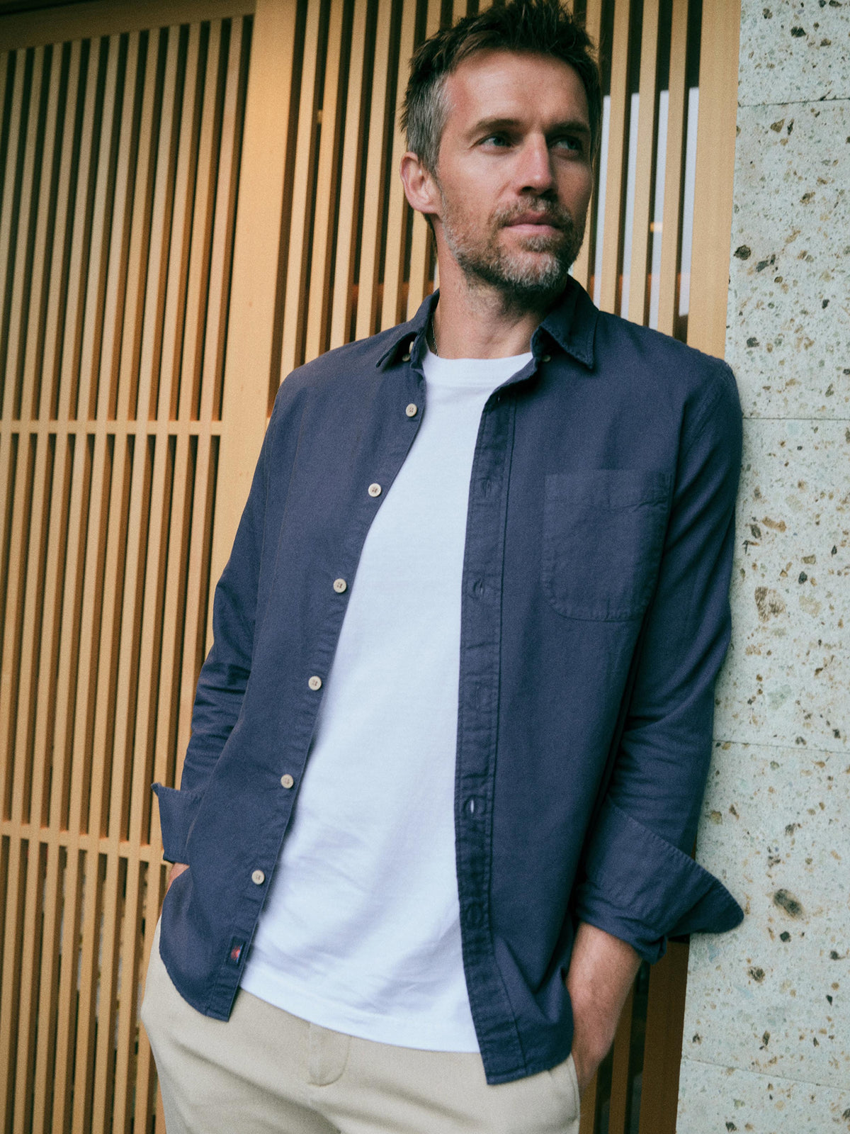 A man wearing a navy blue long-sleeve button-down shirt over a white t-shirt and beige pants, leaning against a modern wooden slat wall and concrete pillar., image 1 of 7