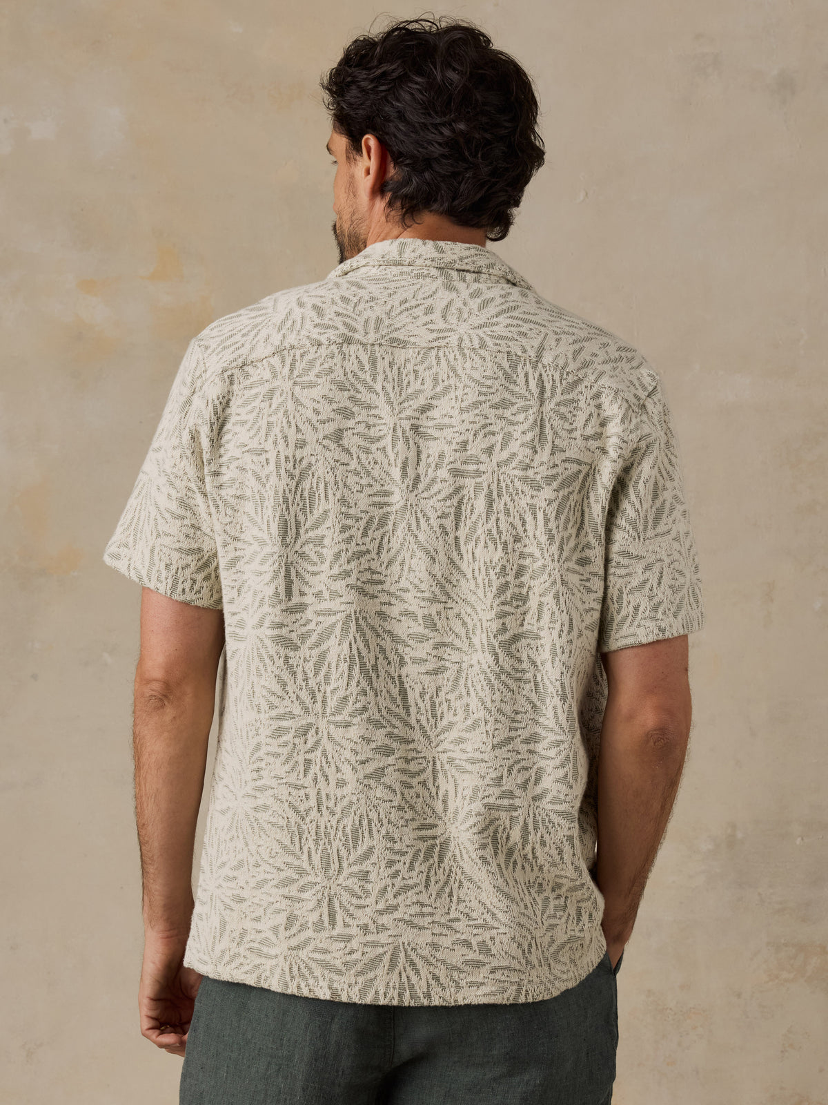 A man viewed from behind wearing a short-sleeve button-up shirt with a light beige and off-white leafy pattern, paired with dark gray trousers, set against a textured beige wall., image 5 of 7