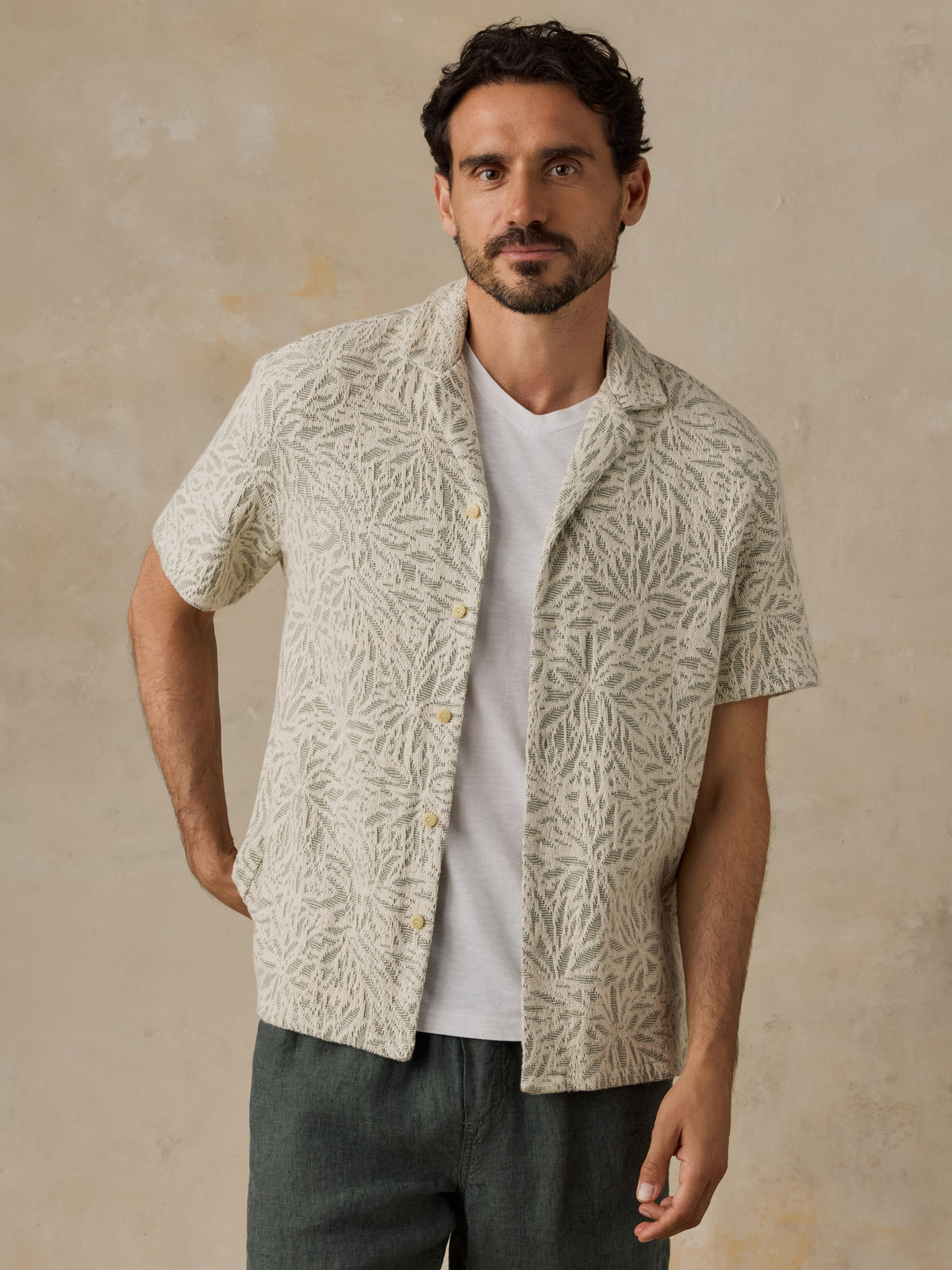 A man wearing a short-sleeve button-front camp shirt with a beige and off-white botanical pattern over a white t-shirt and dark gray trousers, standing against a textured beige wall., image 2 of 7