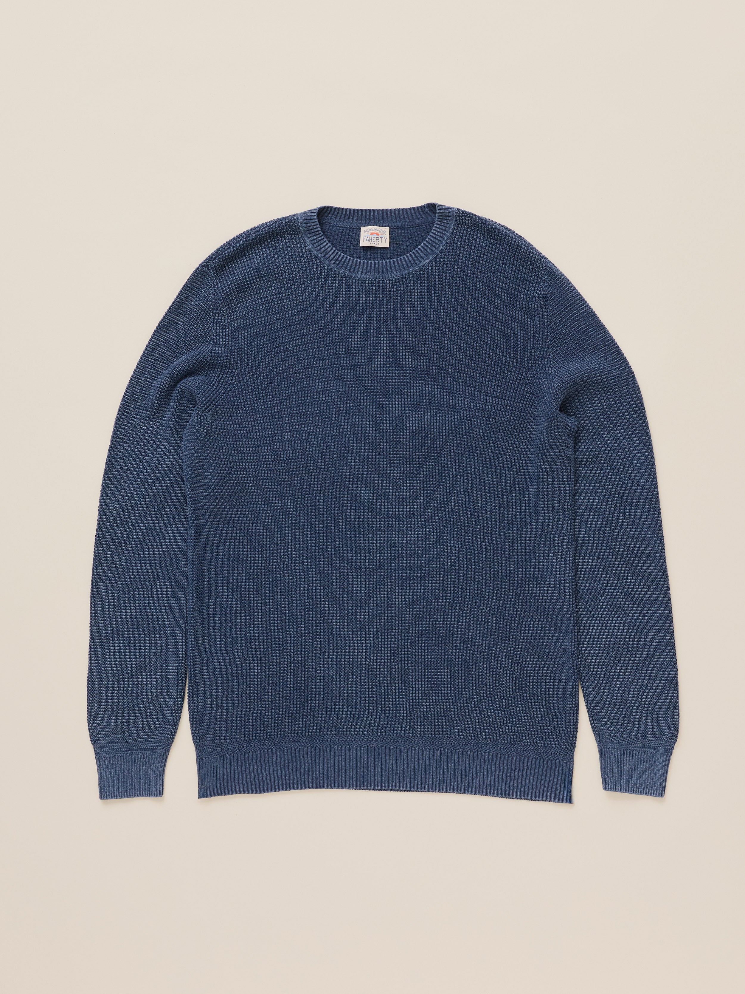 A classic crewneck knit sweater in a muted slate blue color, laid flat against a solid light beige background.