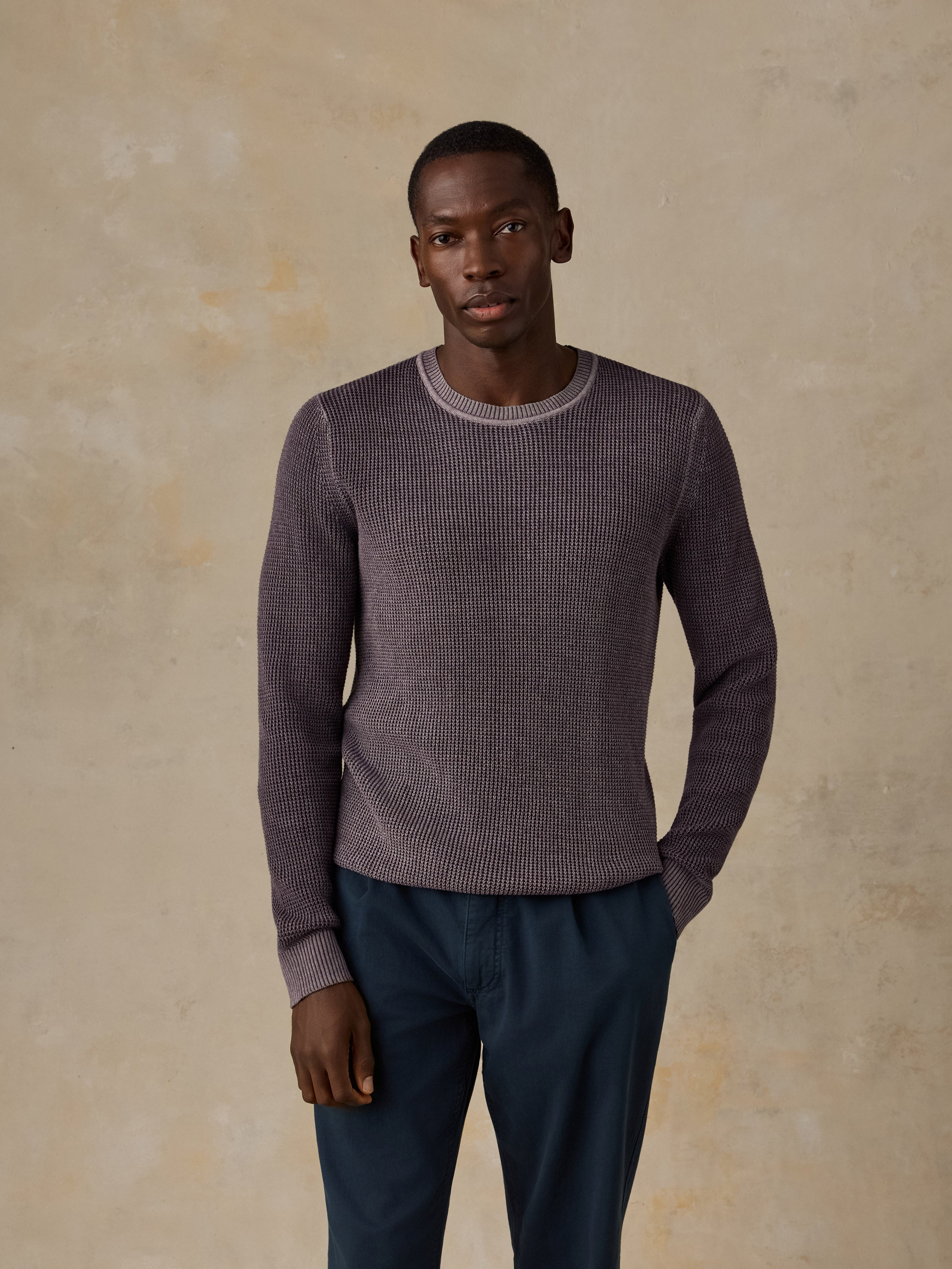 Man wearing a purple knit sweater and navy chino pants against a beige background
