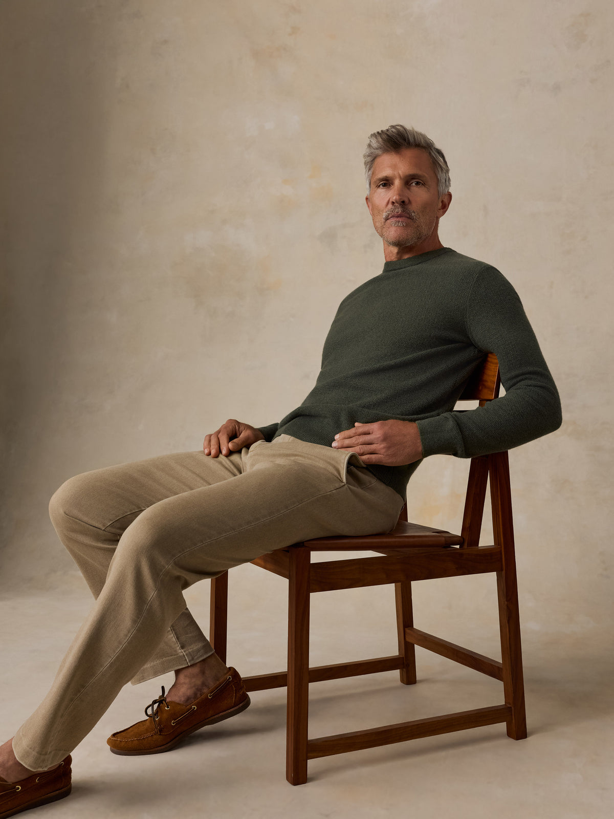 A man sitting on a chair wearing a dark green sweater and beige pants, image 2 of 5