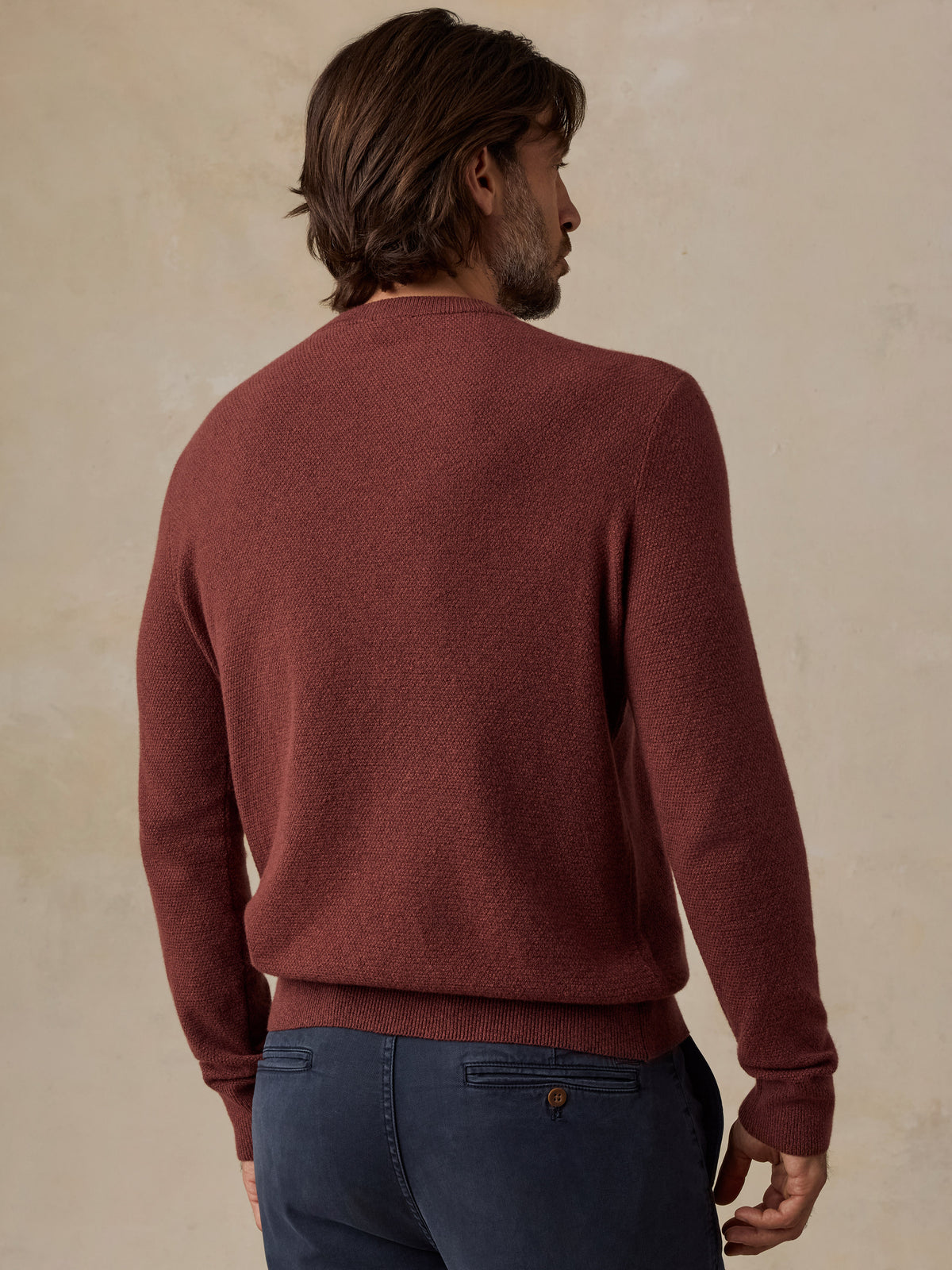 A man viewed from behind wearing a rust-brown long-sleeve knit sweater and dark blue jeans, standing against a textured beige wall., image 2 of 5