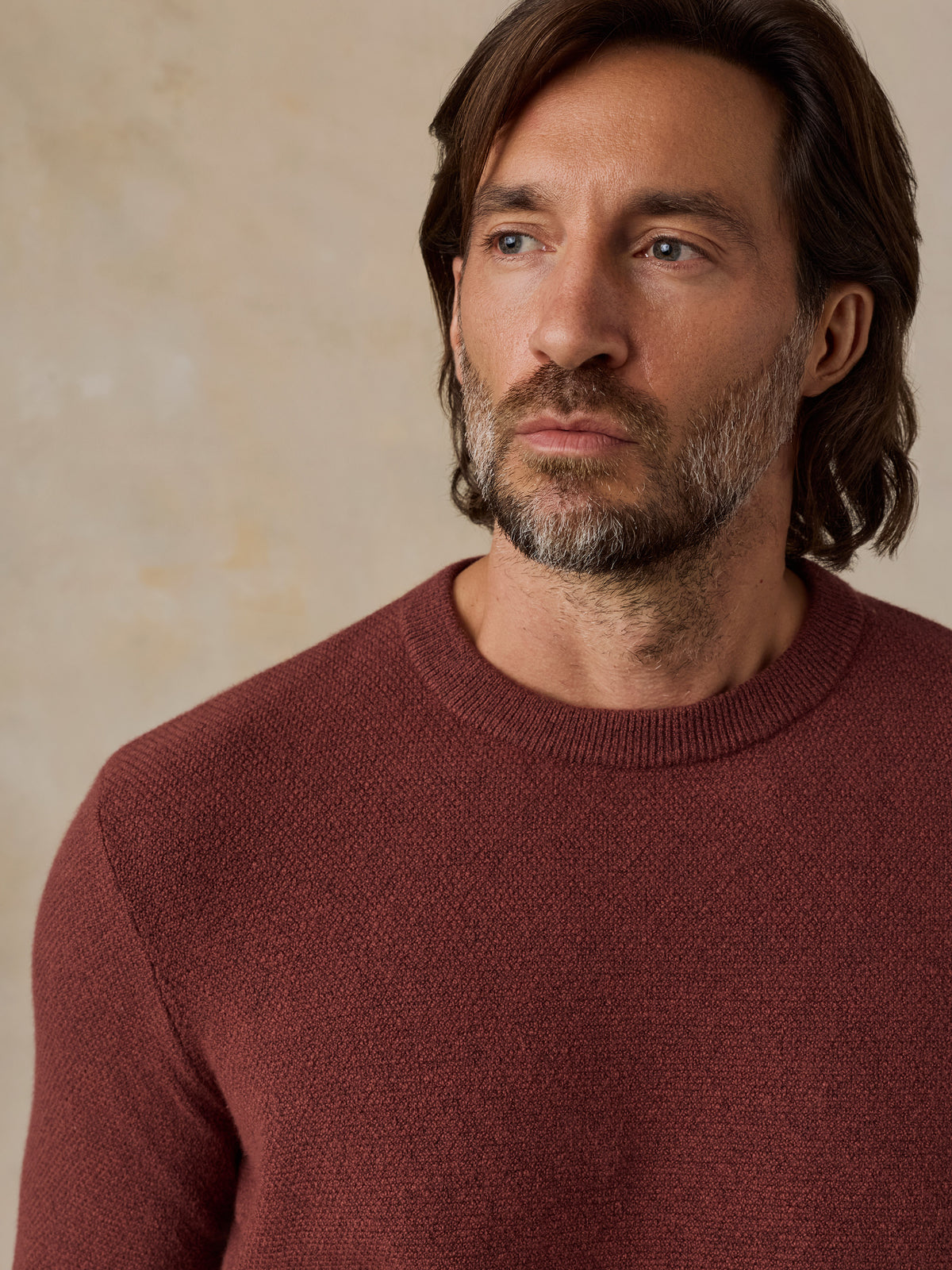 A man wearing a textured crewneck sweater in a muted burgundy or rust color, photographed against a warm beige textured wall., image 3 of 5