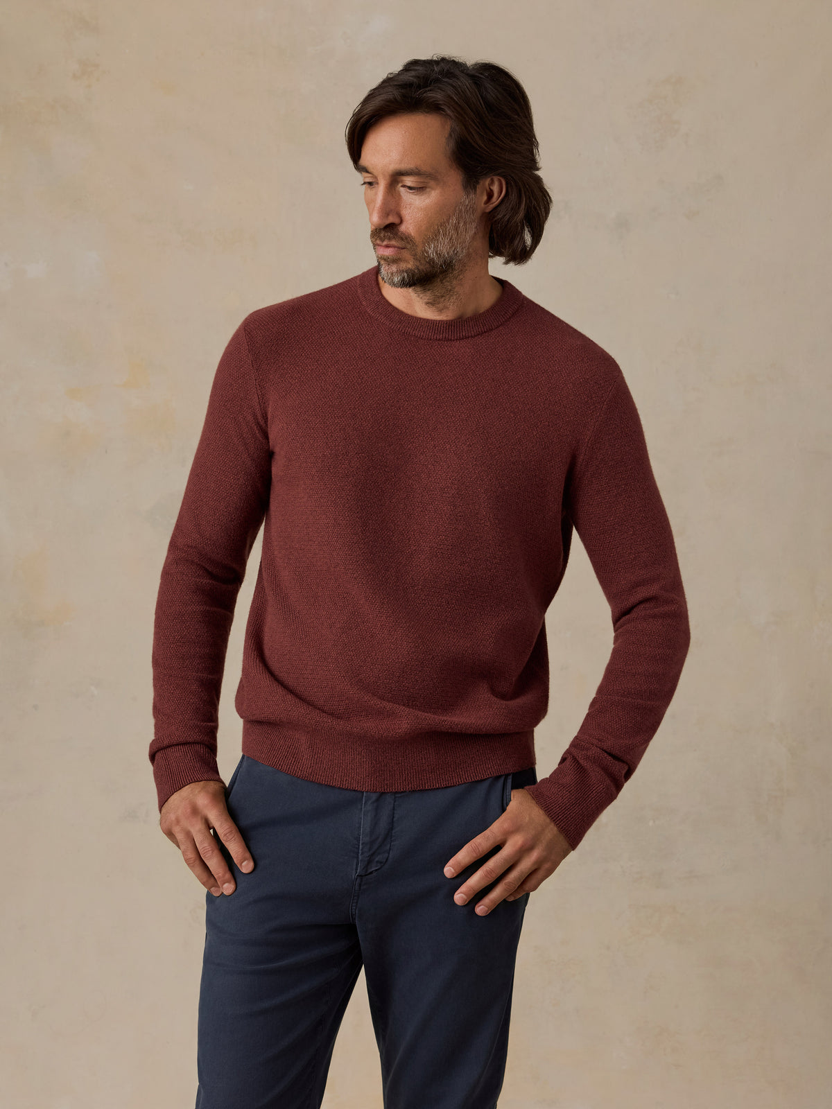 A man wearing a long-sleeve crewneck sweater in a deep burgundy red, paired with navy blue pants, standing against a textured beige studio backdrop., image 4 of 5
