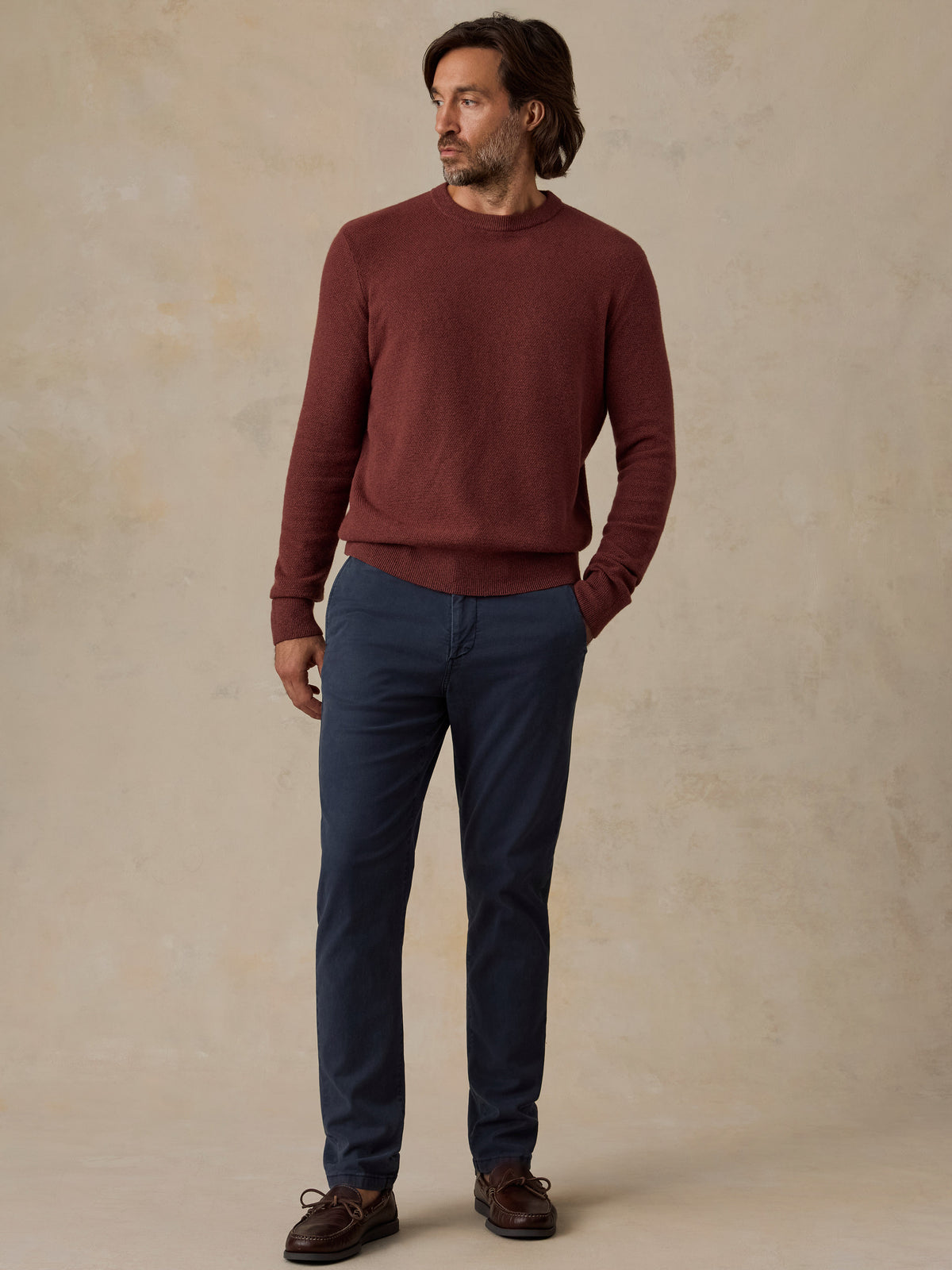 A man wearing a burgundy knit crewneck sweater, navy blue trousers, and brown leather shoes, standing against a textured beige studio backdrop., image 5 of 5