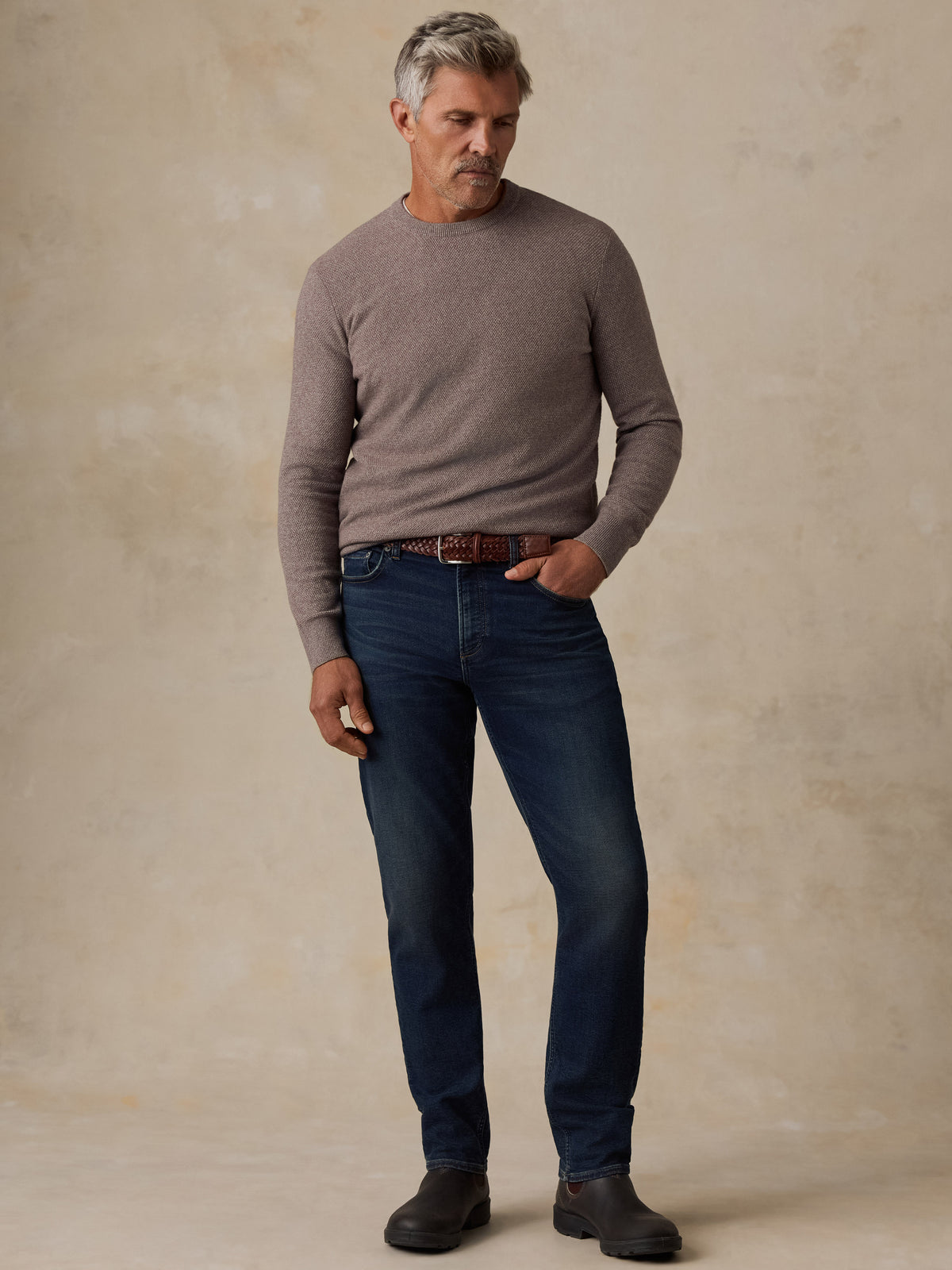 A man wearing a taupe knit crewneck sweater, dark blue jeans, a brown leather belt, and dark brown boots, standing against a textured beige studio backdrop., image 5 of 5