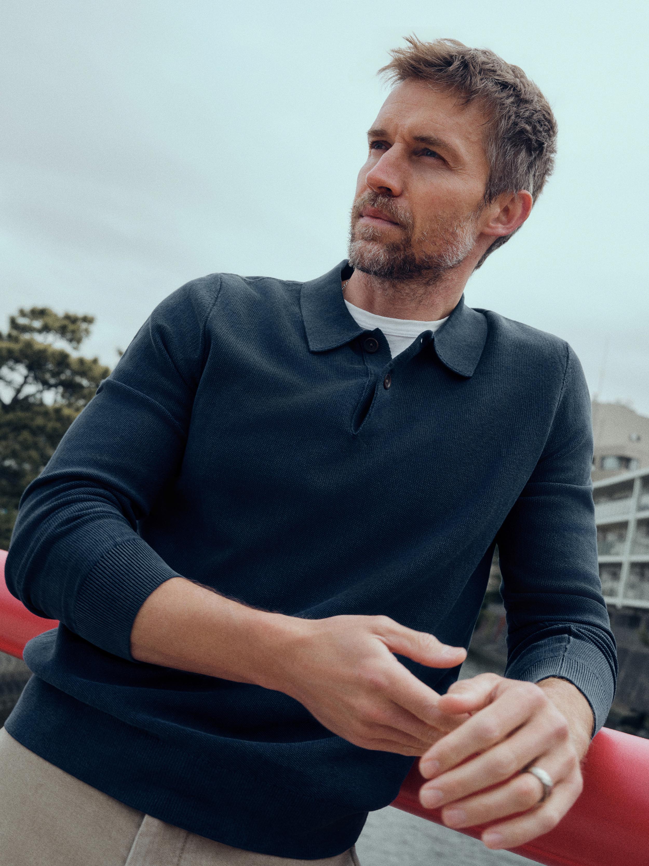 A man wearing a dark navy long-sleeve polo sweater over a white undershirt, leaning against a red railing outdoors under an overcast sky.