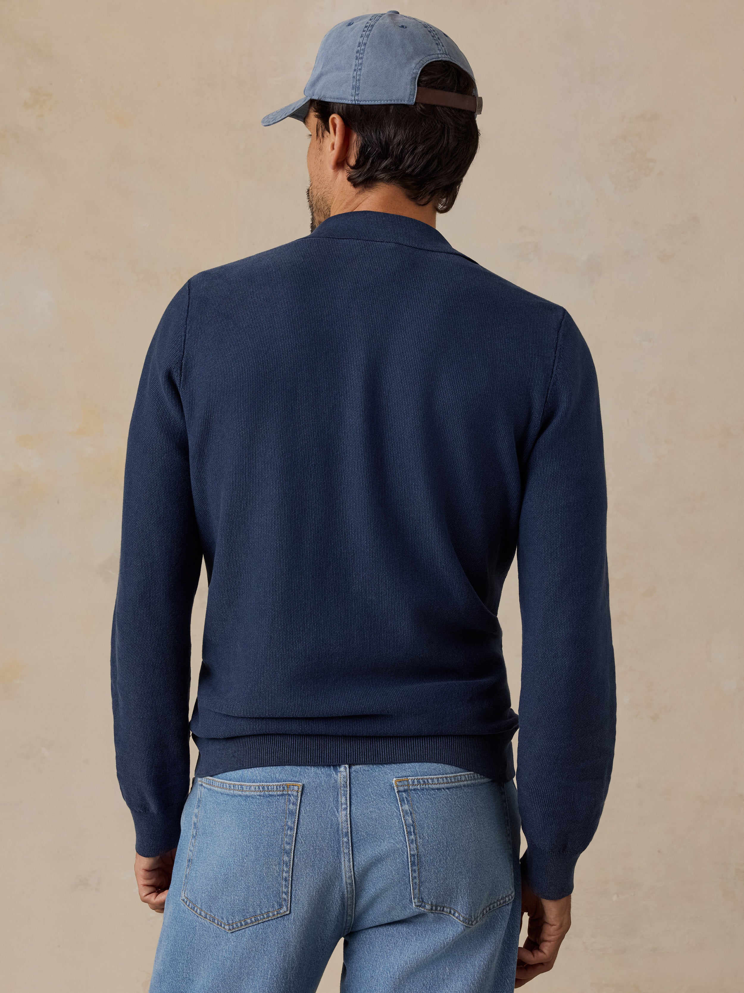 A man viewed from behind wearing a navy blue knit sweater, light blue jeans, and a blue baseball cap against a textured beige background.