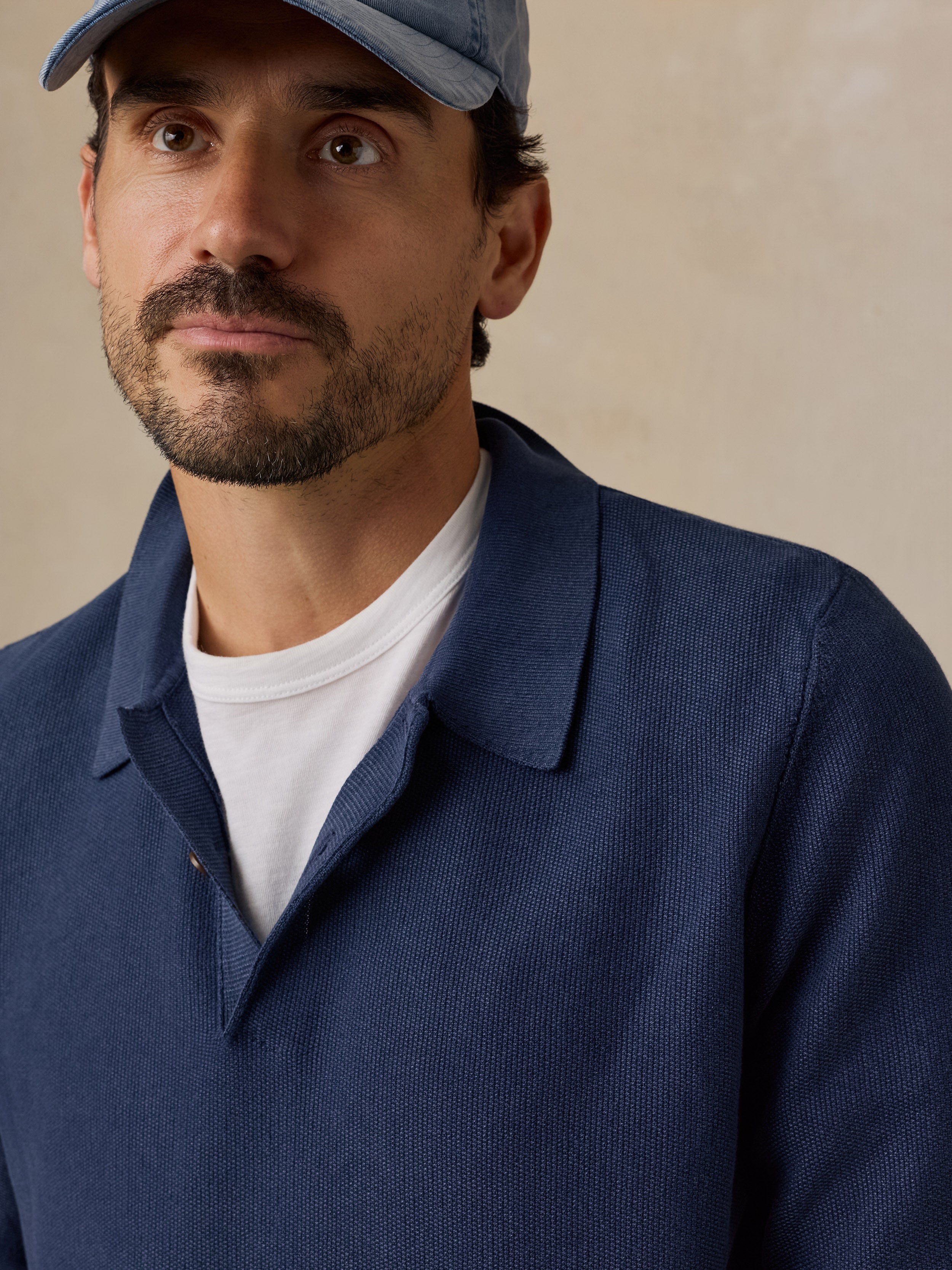 A close-up of a man wearing a textured navy blue polo shirt over a white t-shirt, paired with a light blue baseball cap, set against a neutral beige background.