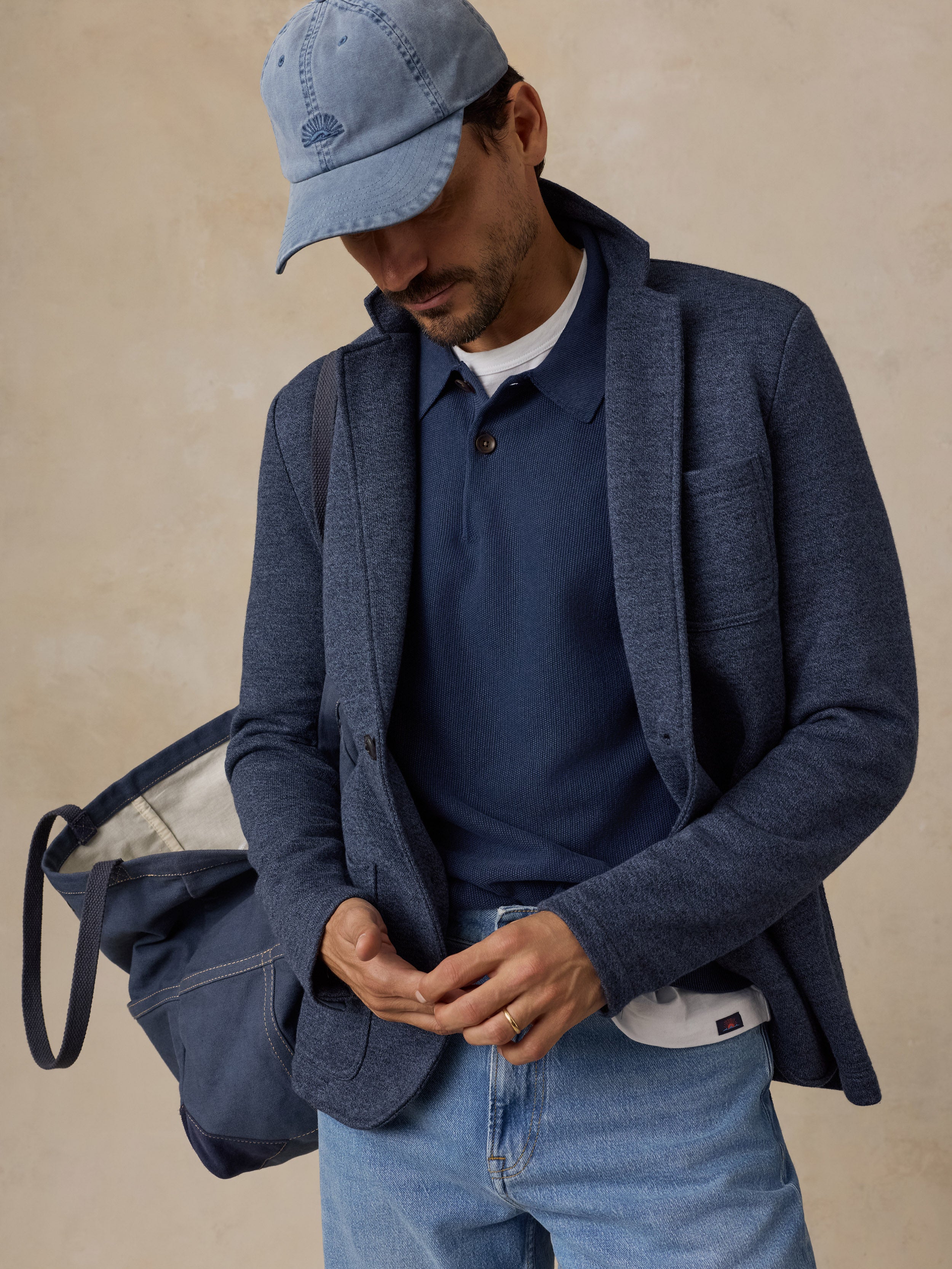 A man wearing a textured navy blue blazer over a navy polo shirt, light blue jeans, and a light blue baseball cap, carrying a navy tote bag against a warm beige background.