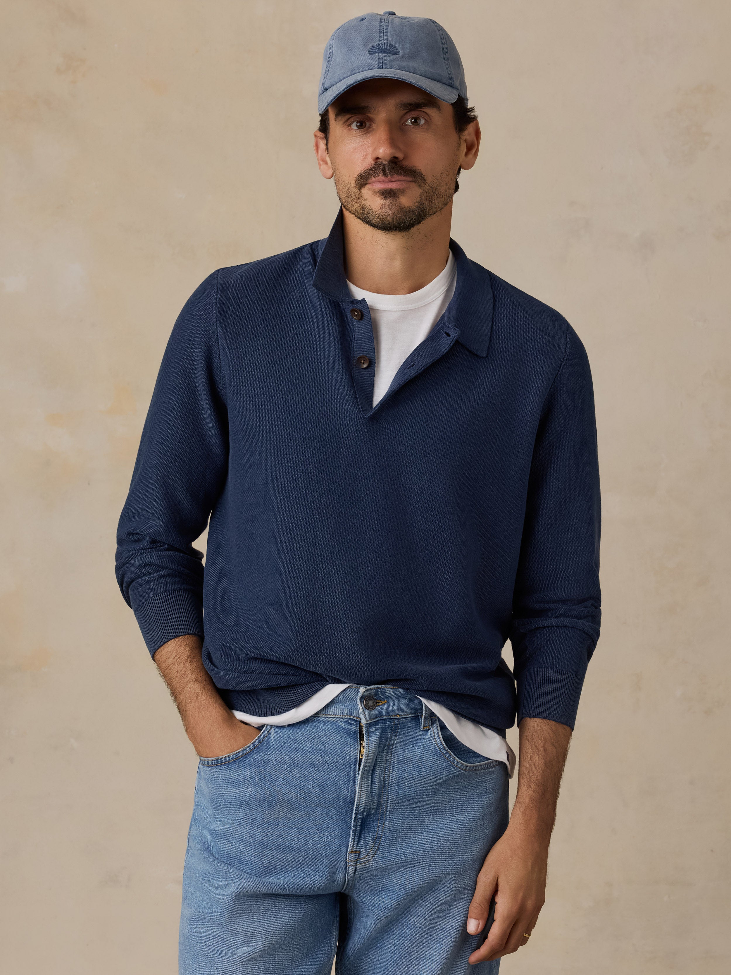 A man wearing a navy blue long-sleeve polo sweater over a white t-shirt, paired with light blue jeans and a matching blue baseball cap, standing against a textured beige background.