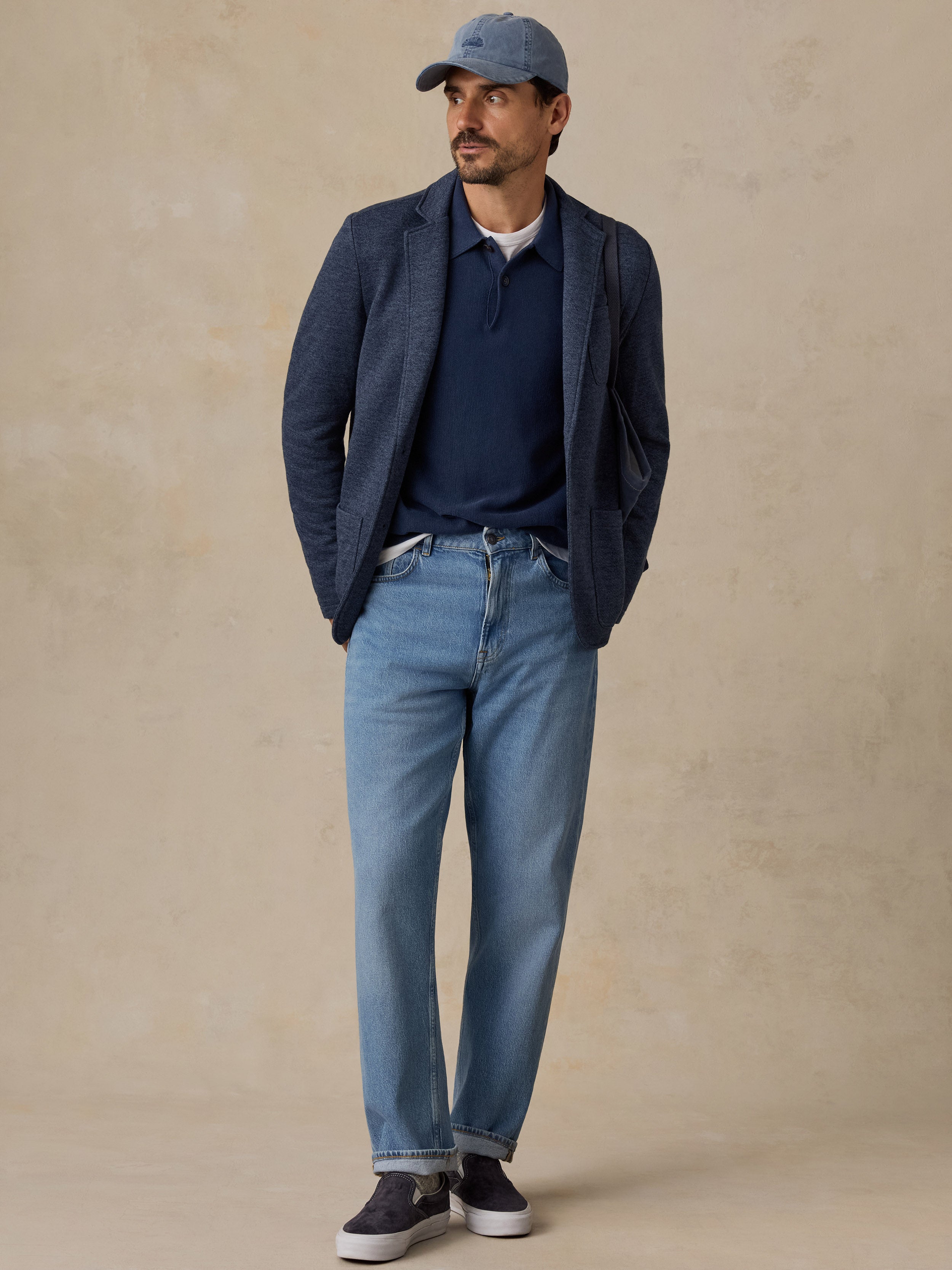 A man wearing a navy blue blazer, navy polo shirt, light blue jeans, a blue baseball cap, and dark sneakers, standing against a textured beige studio backdrop.
