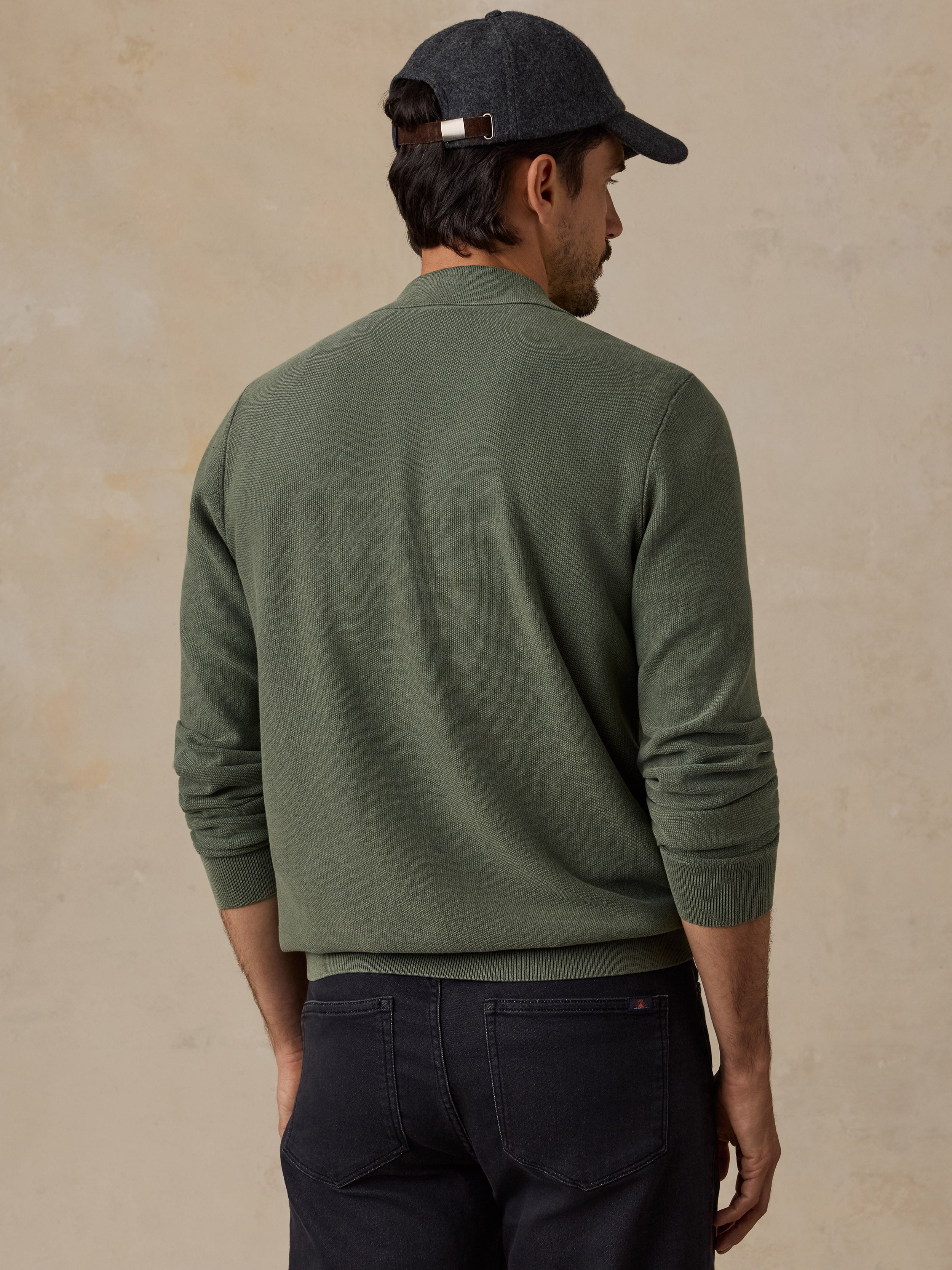 A man shown from the back wearing an olive green knit sweater, dark navy pants, and a dark gray baseball cap, set against a textured beige background.