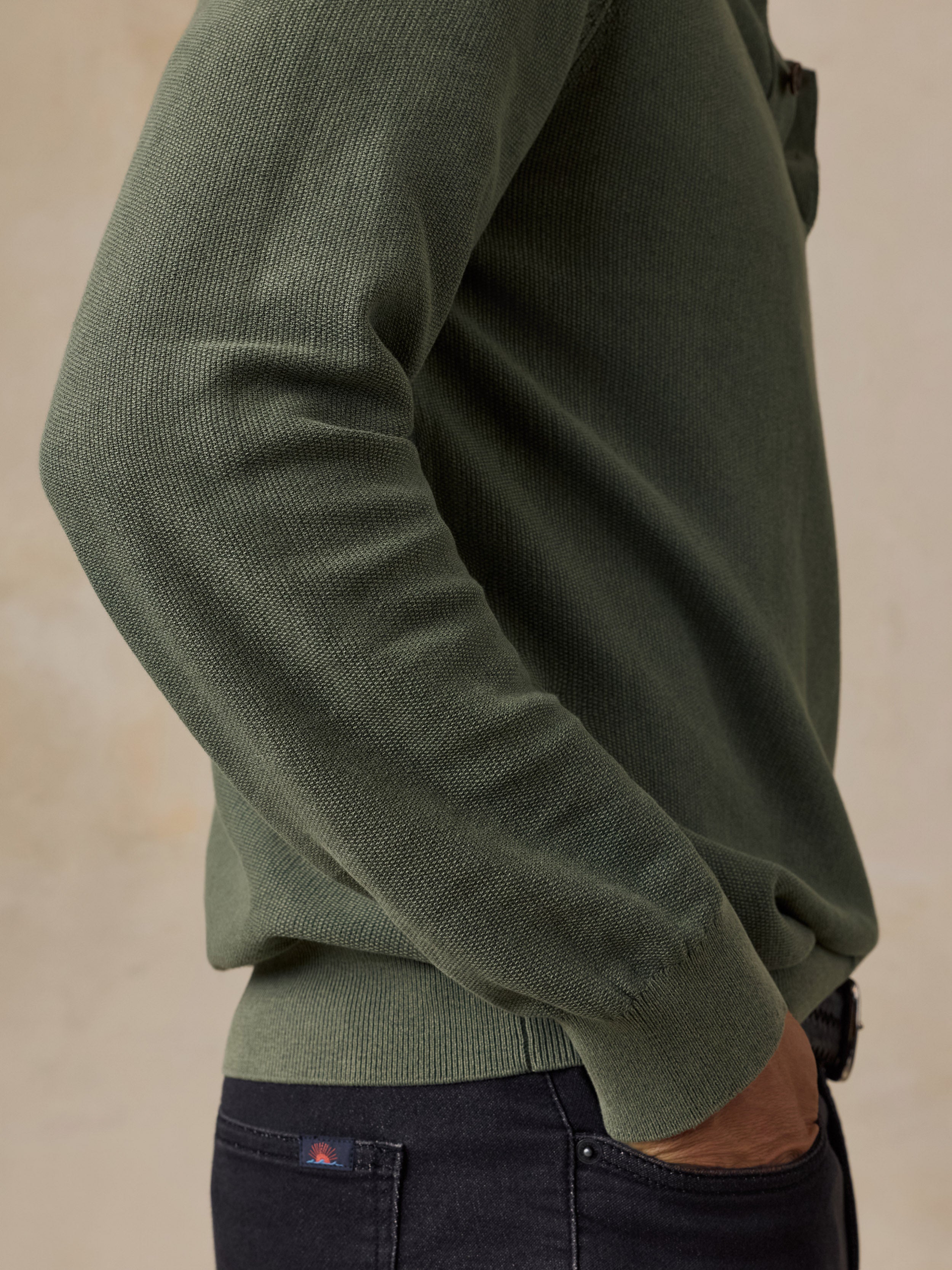 A close-up side view of a man wearing an olive green ribbed knit sweater and dark navy jeans against a warm beige background.
