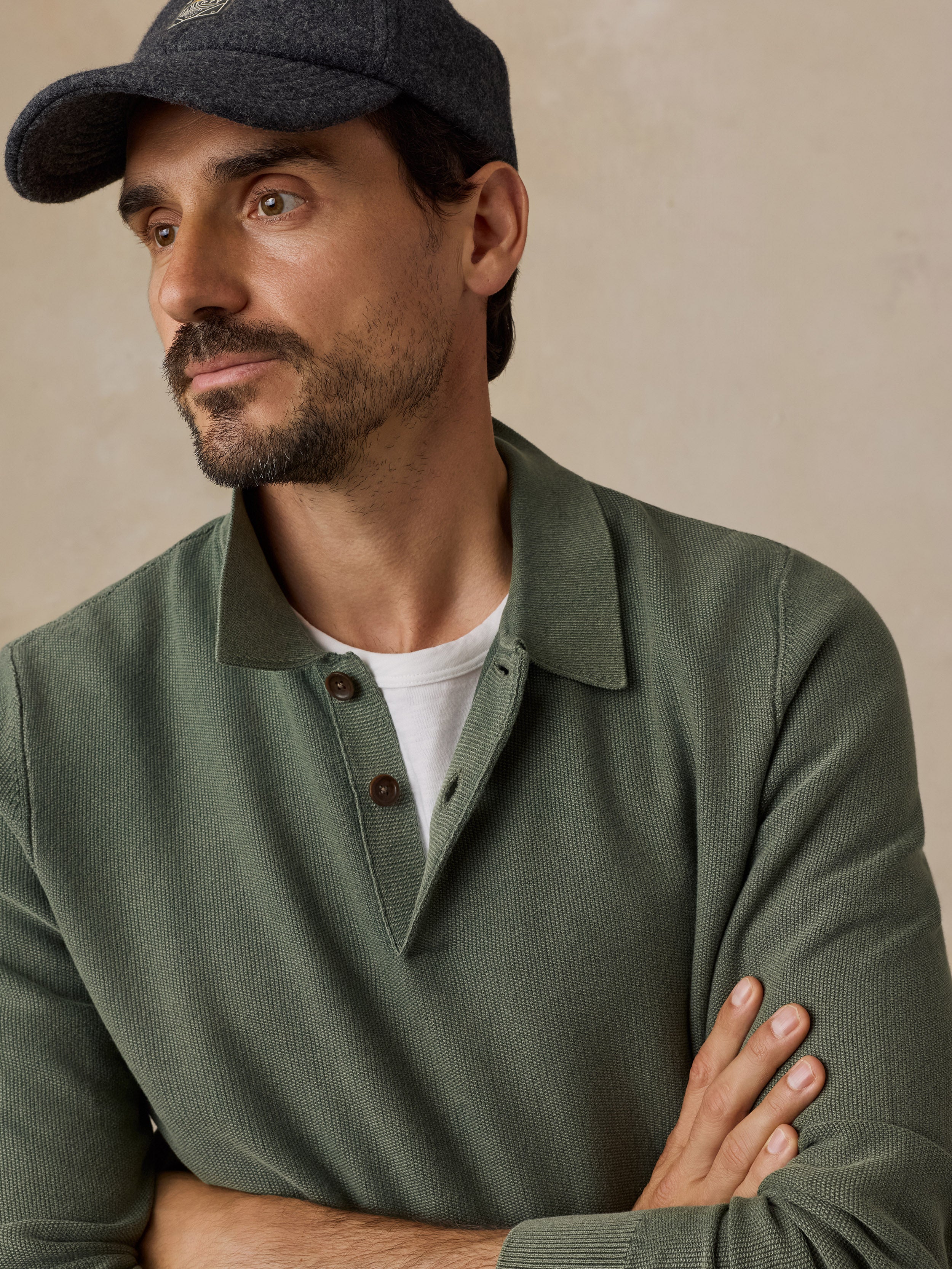 A man wearing an olive green knit polo henley sweater over a white t-shirt, paired with a dark gray baseball hat, set against a warm beige textured background.