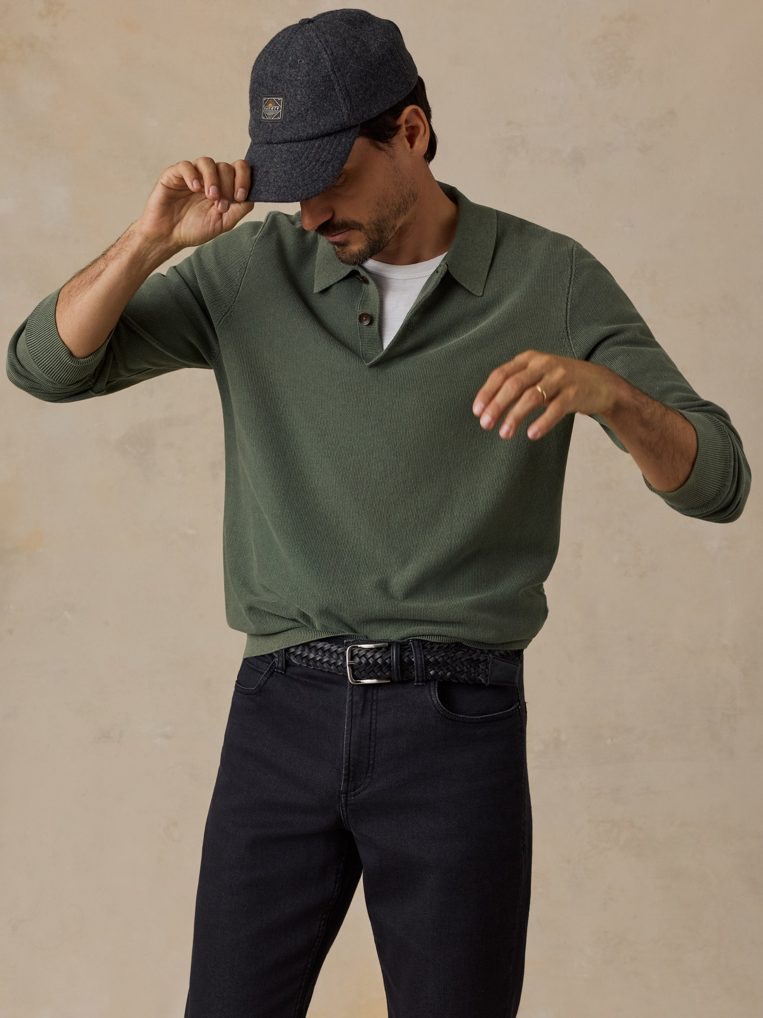 A man wearing an olive green long-sleeve polo shirt, dark navy pants, a dark grey cap, and a white undershirt, posing against a textured beige background.