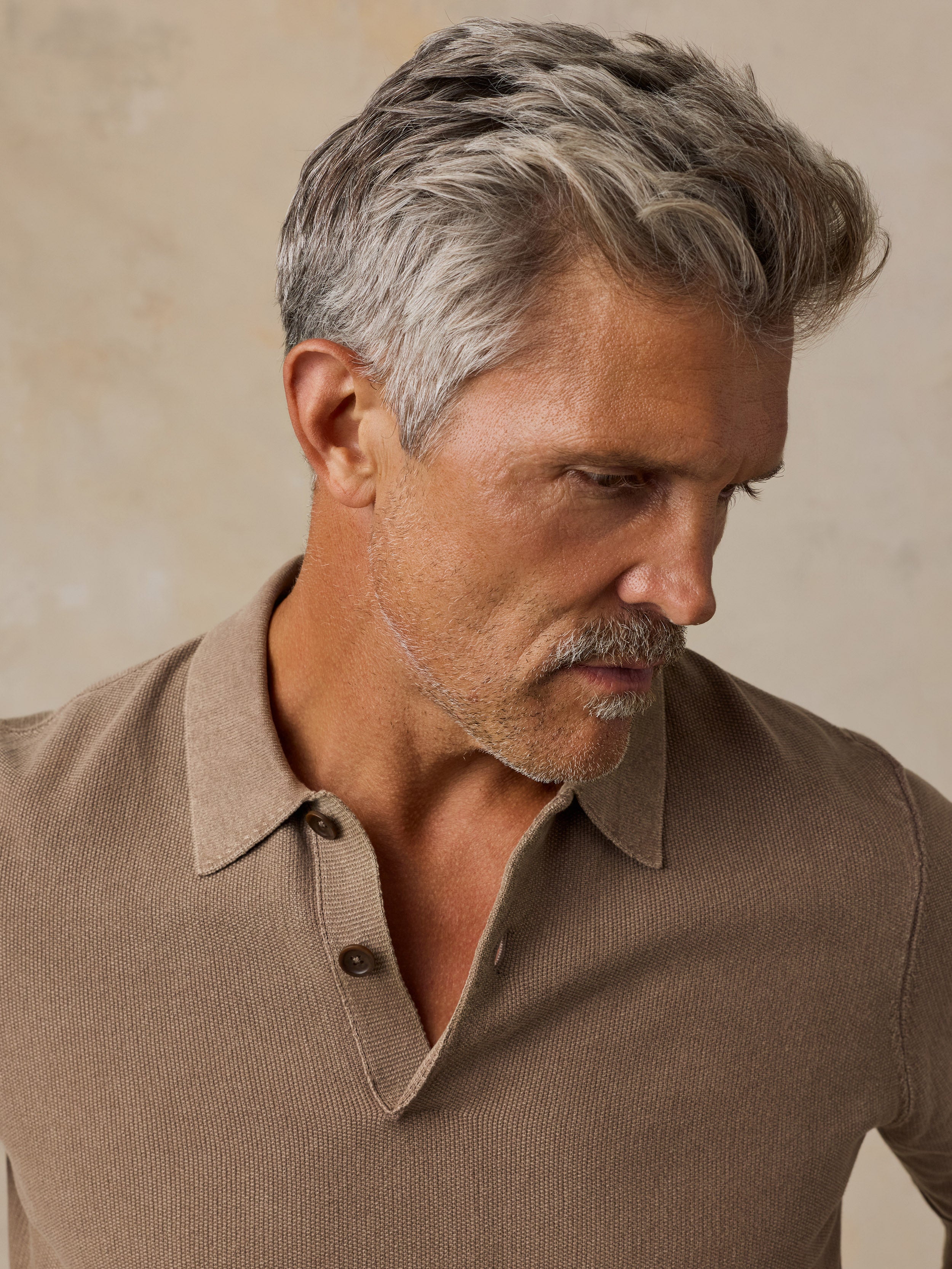 A close-up of a man wearing a brown ribbed knit polo shirt, looking down and to the side against a textured beige background.