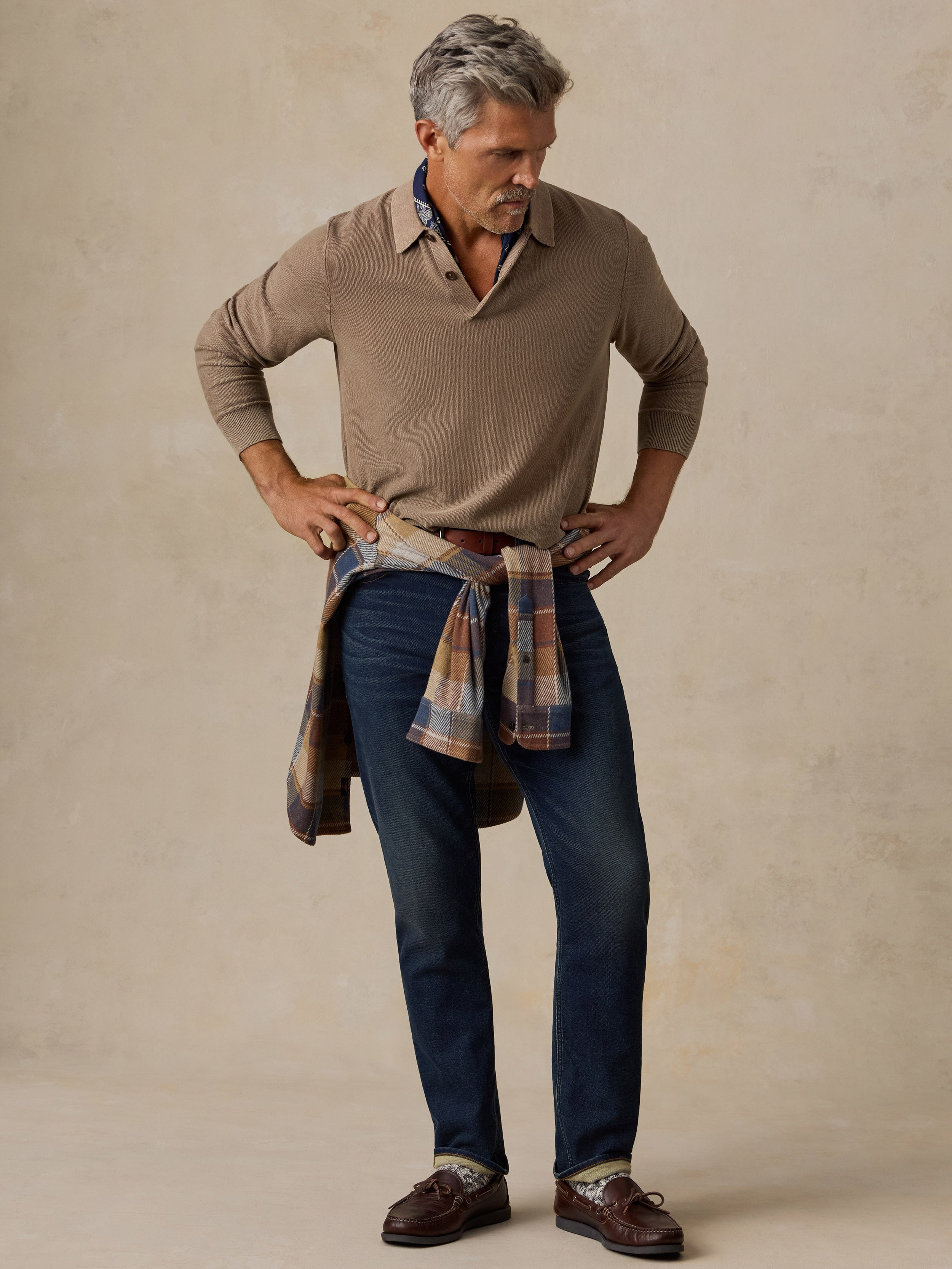 A man modeling a brown long-sleeve knit polo shirt, dark blue jeans, and brown leather shoes, with a plaid shirt tied around his waist, against a textured beige studio backdrop.