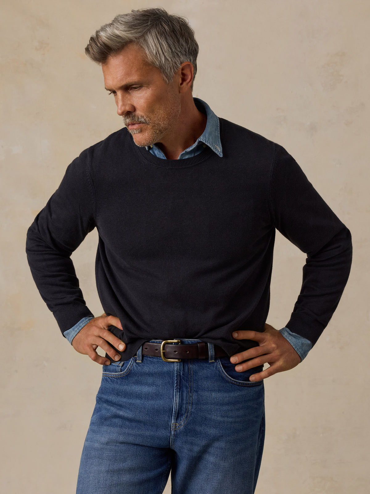 A man wearing a navy long-sleeve knit sweater over a light blue collared shirt, paired with blue jeans and a brown belt.