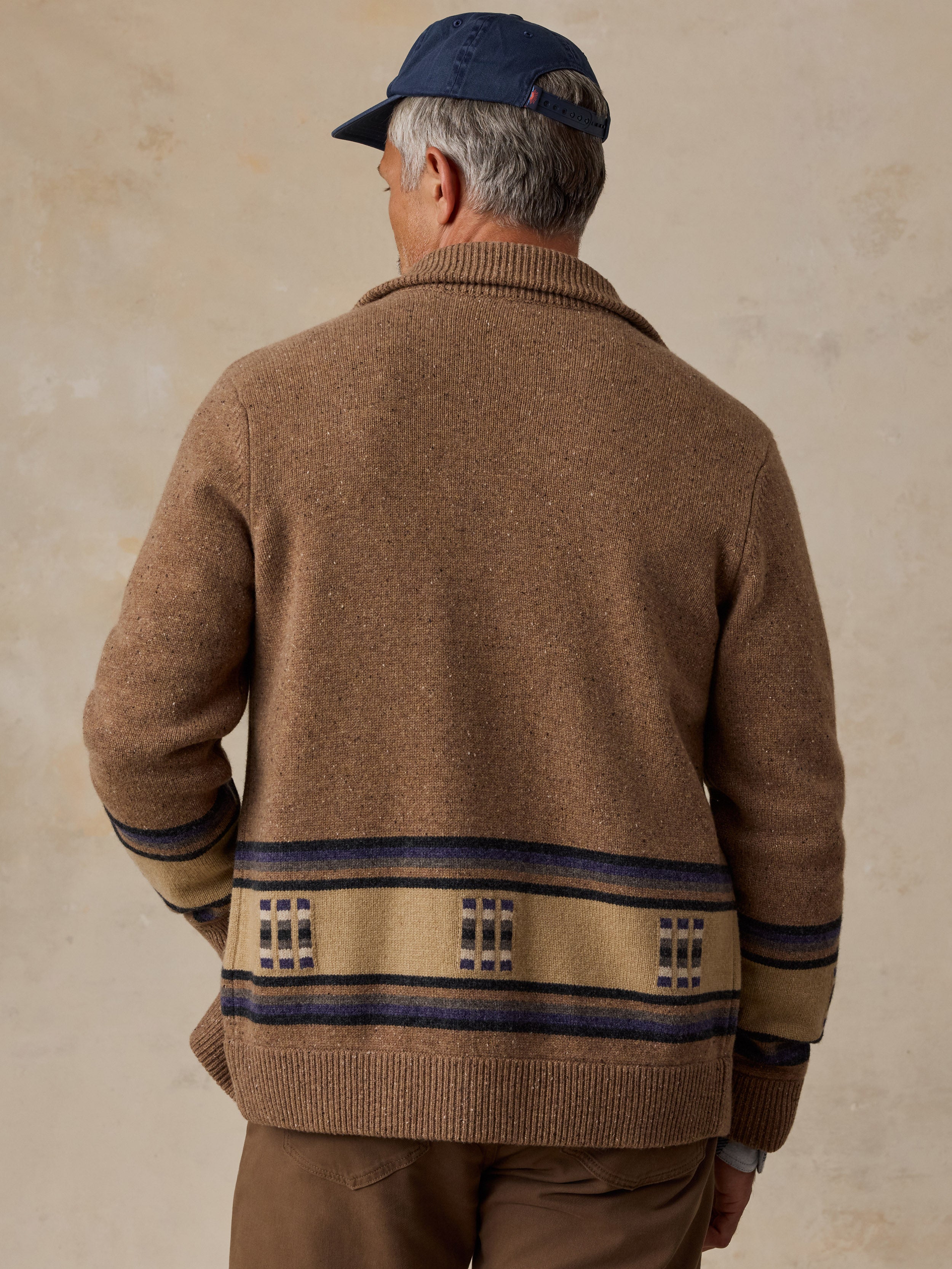 A man viewed from behind wearing a brown knit cardigan with a beige and navy geometric pattern band around the hem and cuffs, paired with brown pants and a navy baseball cap, against a textured beige background.