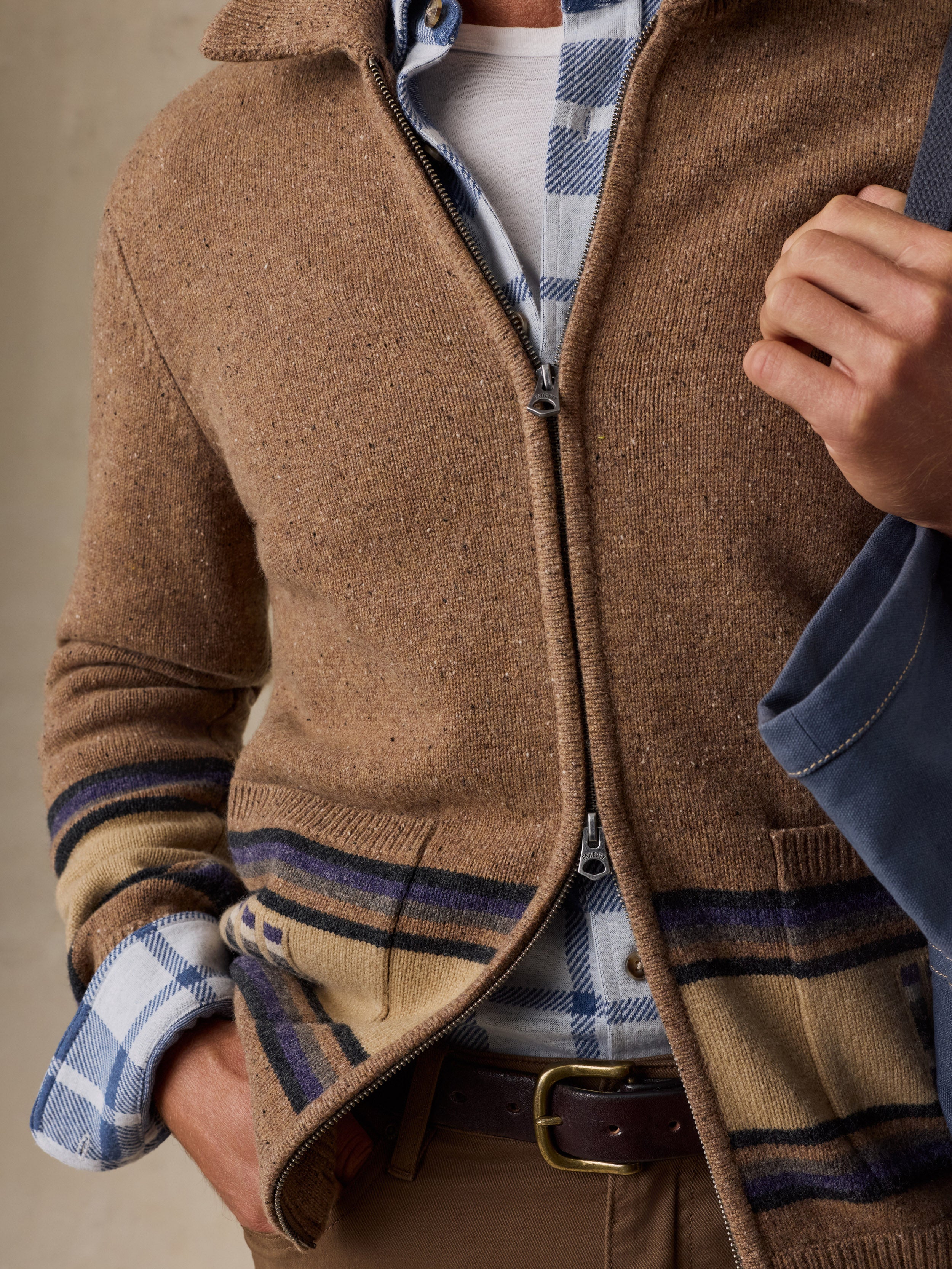 A close-up of a man wearing a brown zip-front knit cardigan with plaid trim, layered over a blue and white plaid shirt and brown trousers.