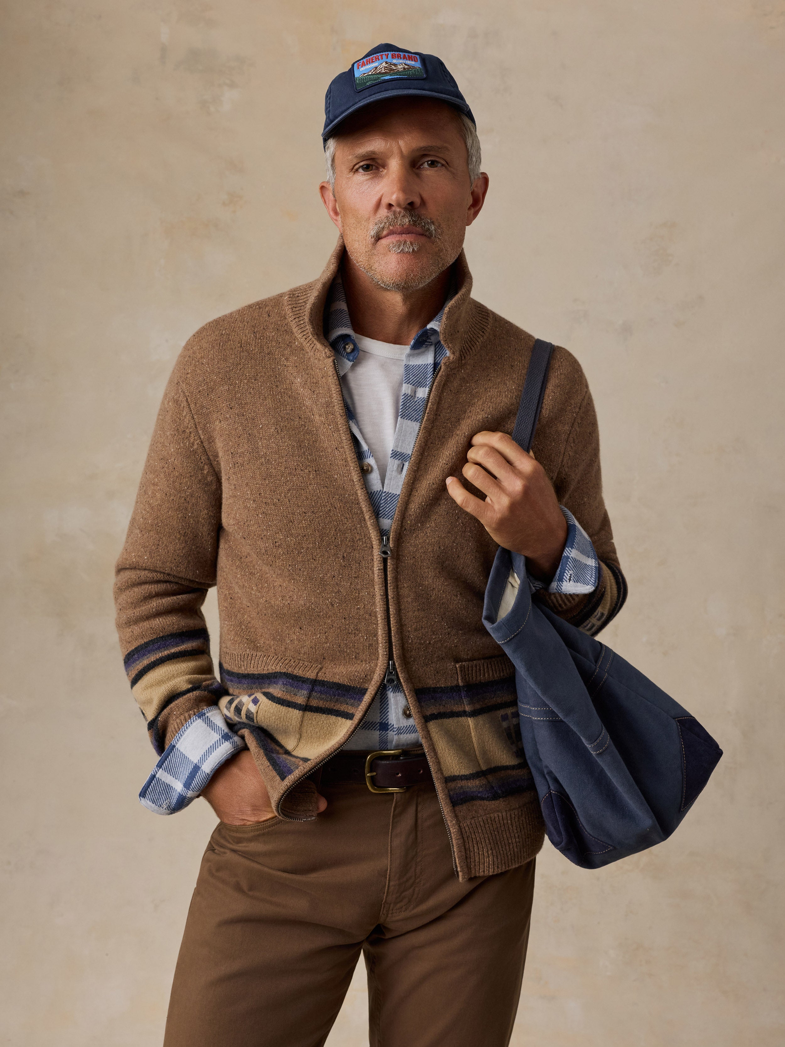 A man wearing a brown zip-up cardigan with a plaid hem, brown pants, a blue plaid button-down shirt, a navy baseball cap, and carrying a navy blue tote bag, posed against a textured beige background.