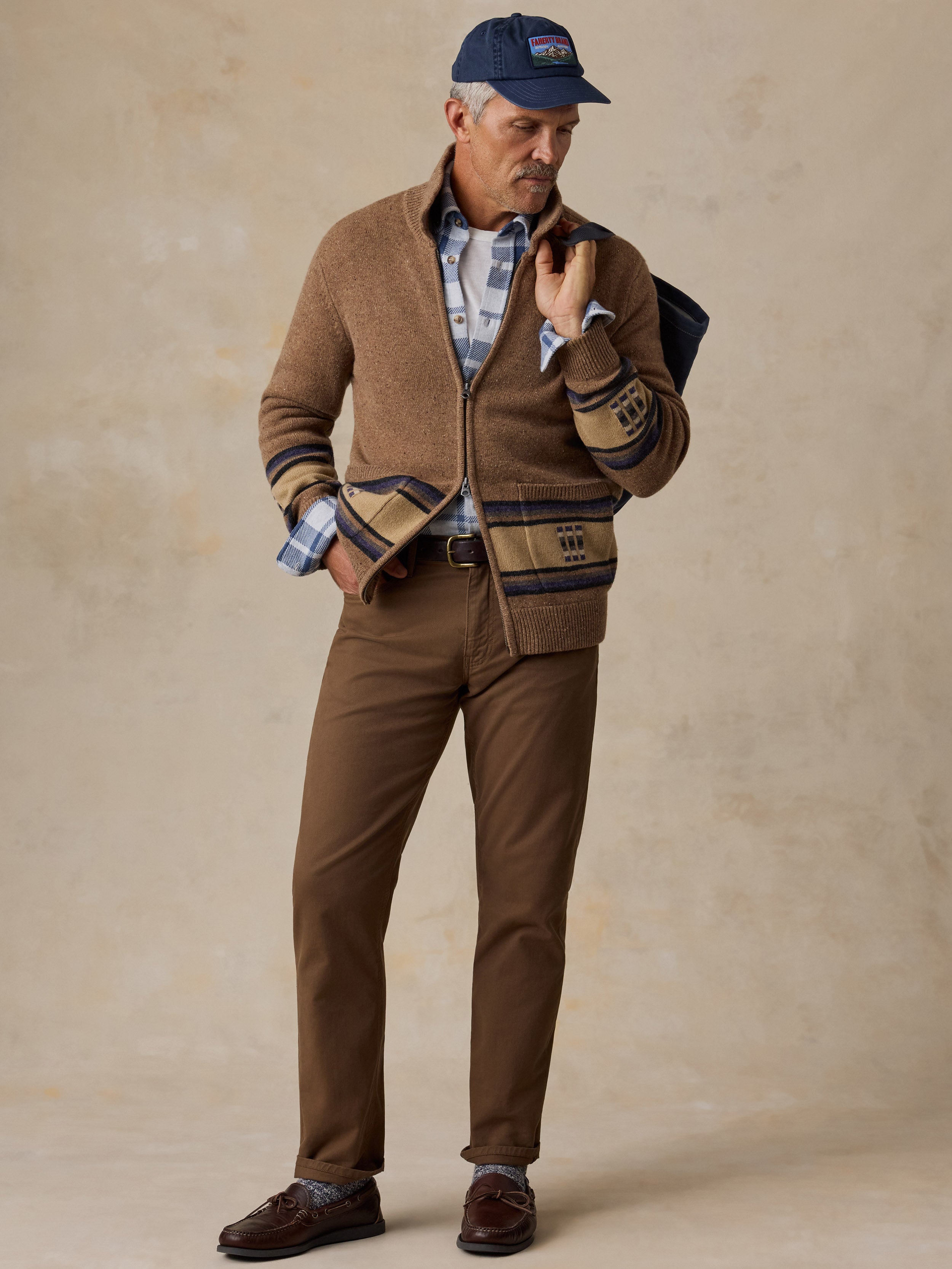 A man modeling a brown knit zip-up cardigan with striped cuffs and hem, paired with brown trousers, a blue plaid shirt, a navy baseball cap, and brown leather shoes against a textured beige studio backdrop.
