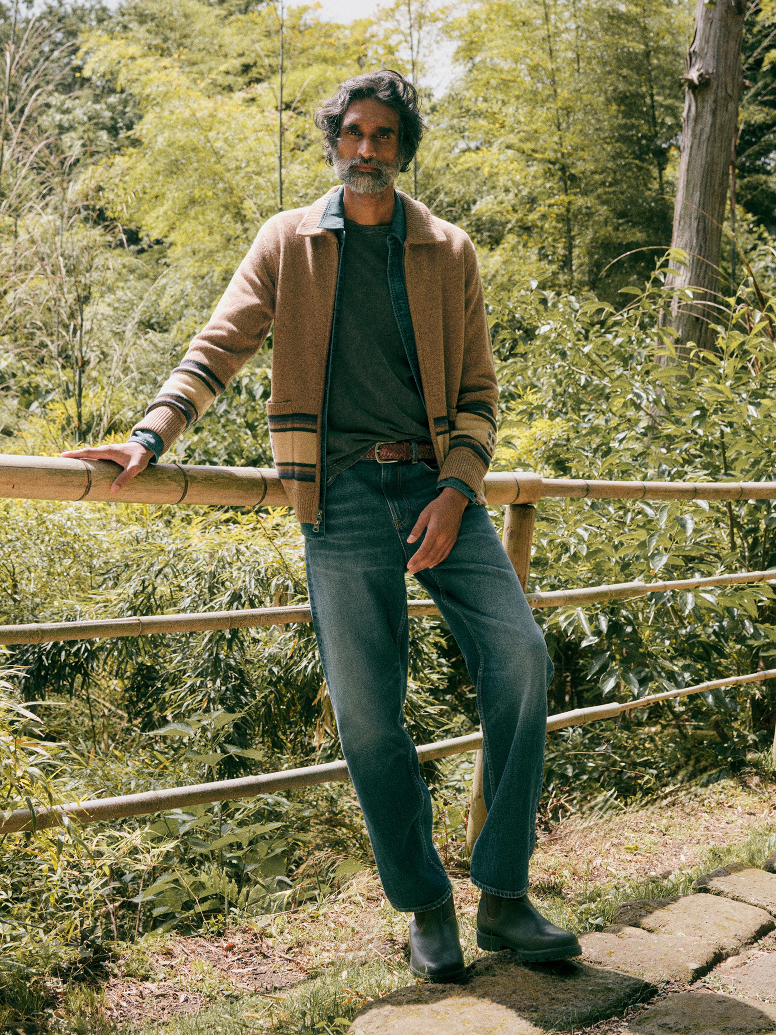 A man wearing a tan corduroy jacket, dark green t-shirt, blue jeans, and dark boots leans against a bamboo fence in a lush green outdoor setting.