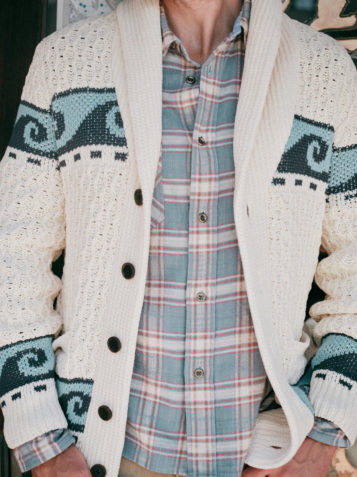 A close-up of a man wearing a cream and blue printed cardigan over a plaid shirt, image 7 of 7