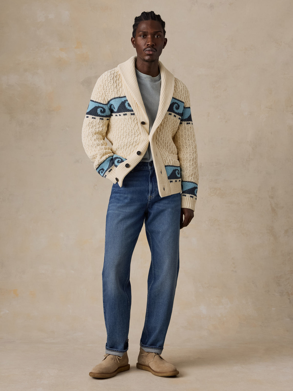 A man wearing a cream and blue printed cardigan with blue jeans and beige boots, image 6 of 7