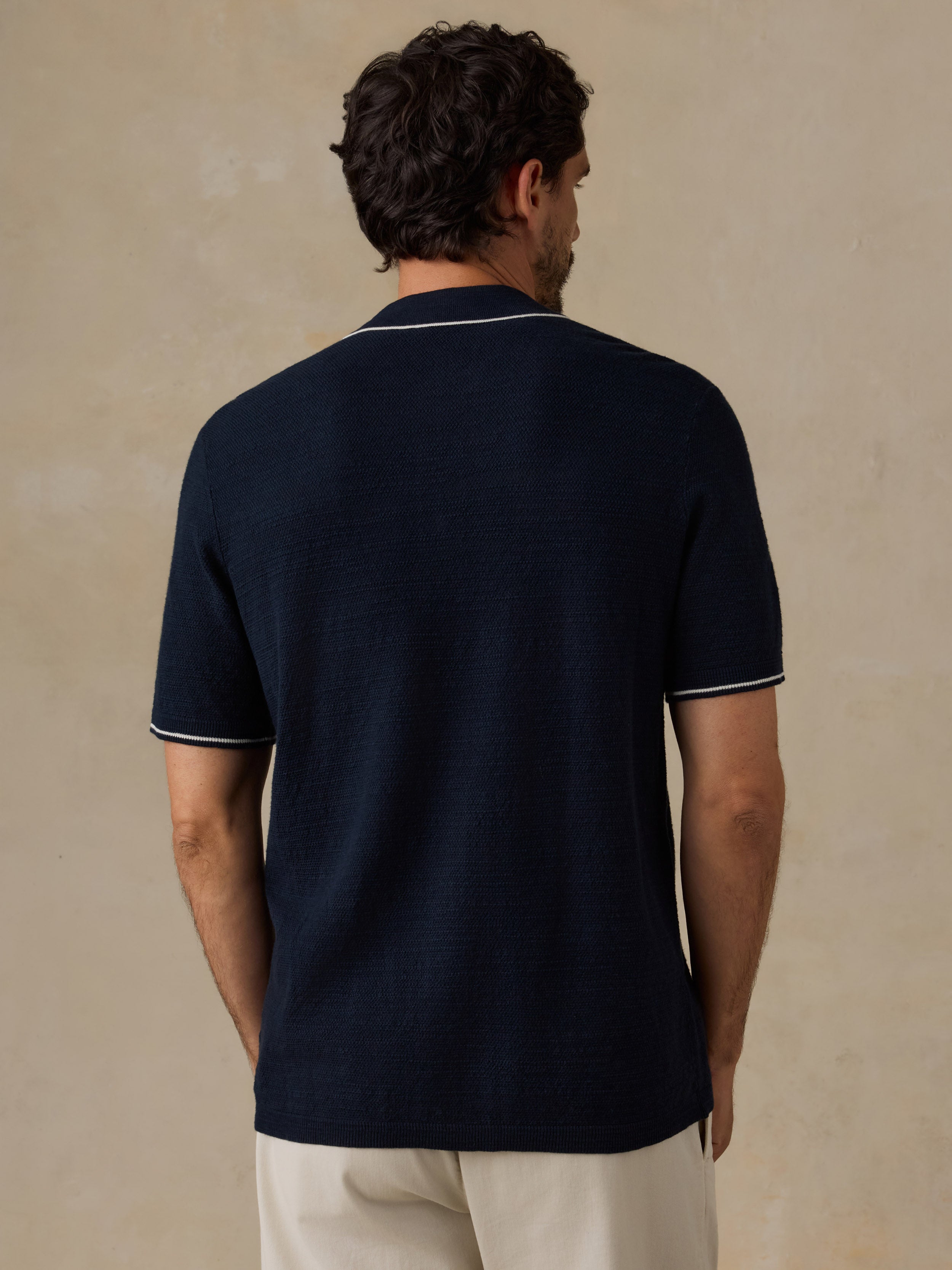 A man viewed from behind wearing a dark navy blue short-sleeve knit polo shirt with thin white piping trim on the collar and sleeve cuffs, paired with off-white trousers, set against a textured beige background.