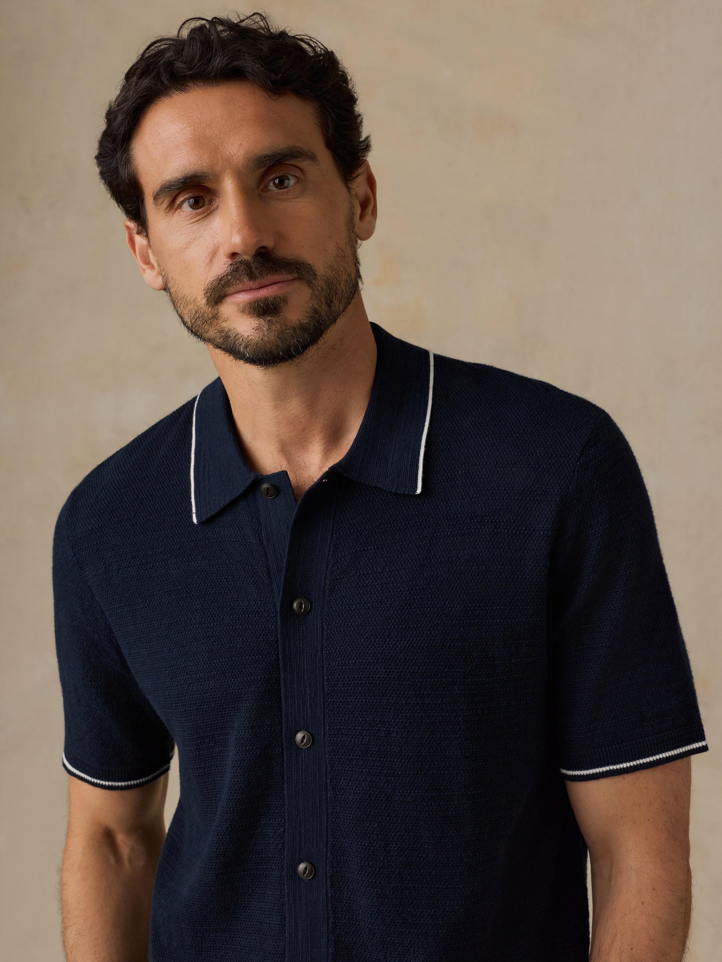 A man wearing a dark navy blue short-sleeve knit polo shirt with white piping on the collar and sleeve cuffs, set against a neutral beige background.